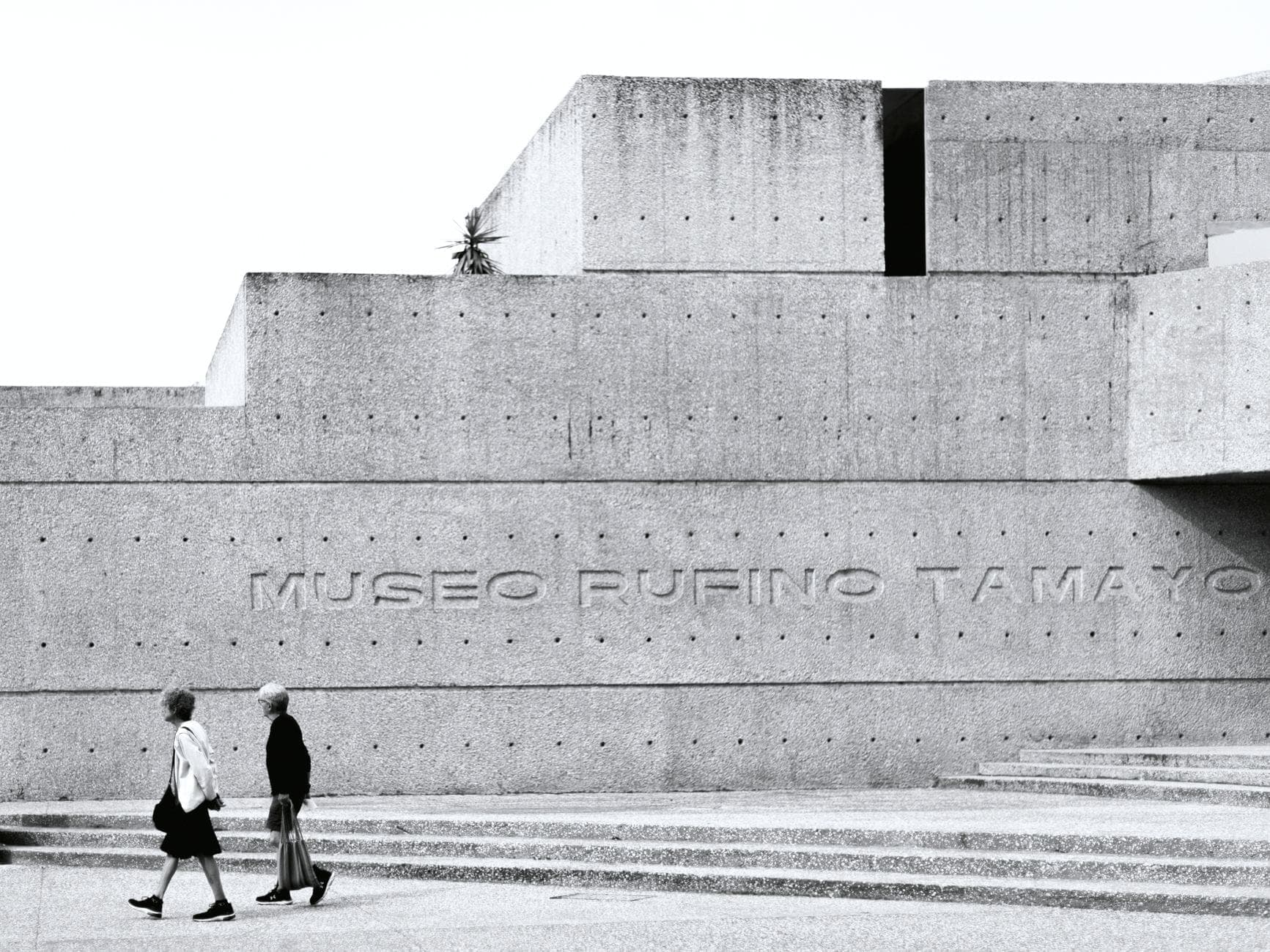 Fachada moderna de concreto del Museo Rufino Tamayo con dos personas caminando frente a él, mostrando el exterior de un reconocido museo de Ciudad de México.