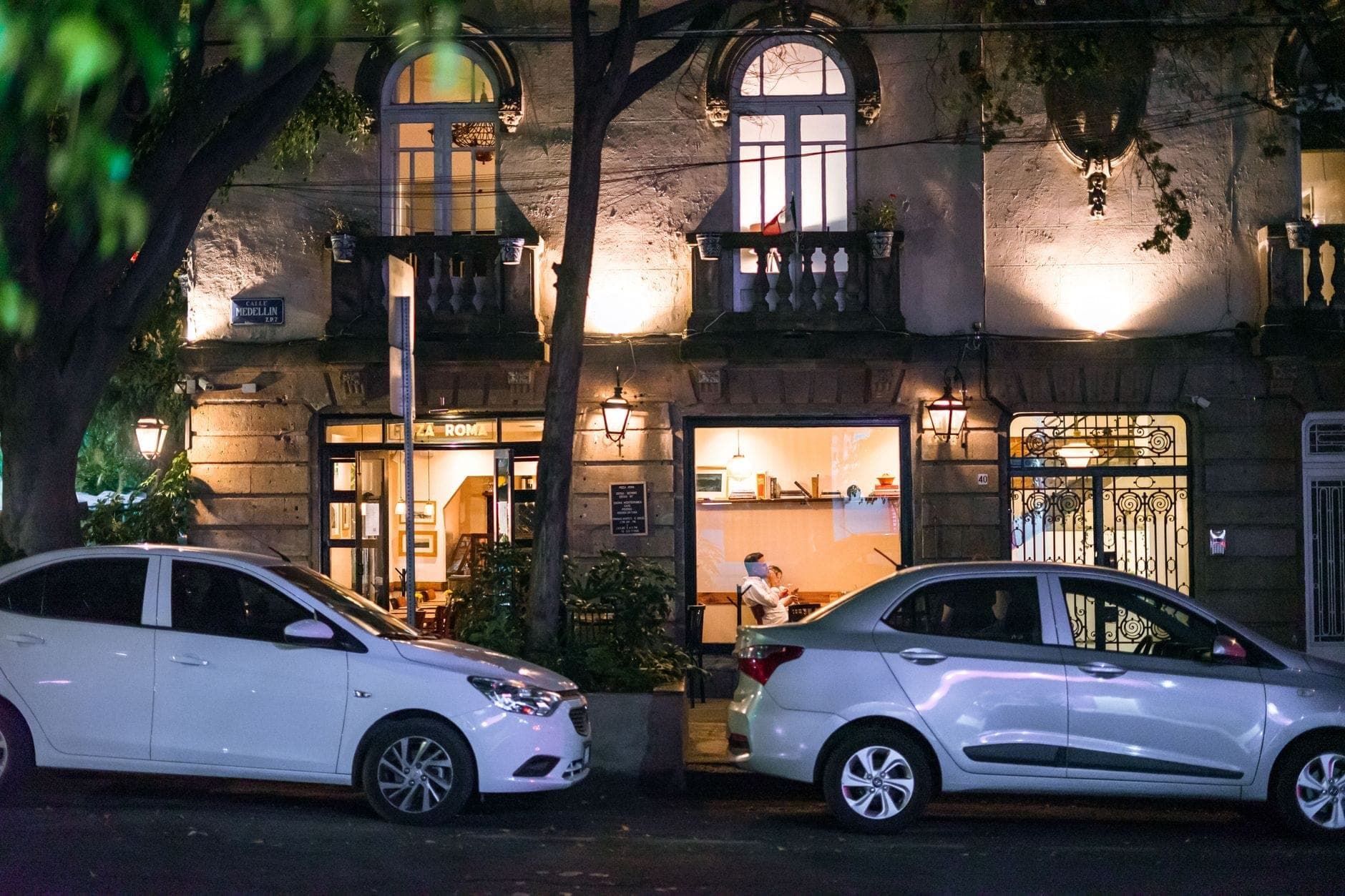 Vista nocturna de un edificio histórico en Roma o Condesa con iluminación estilo boutique, grandes ventanas y coches aparcados en frente.