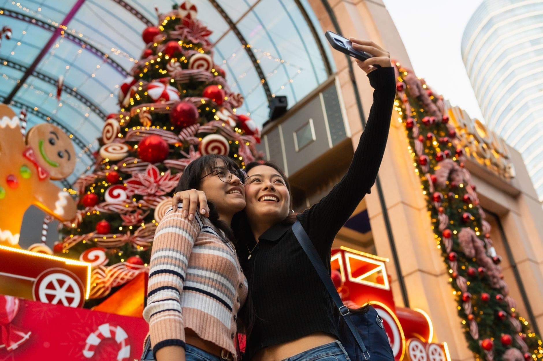 Dos mujeres tomándose una selfie frente a un centro comercial decorado con adornos navideños y una gran exhibición festiva.
