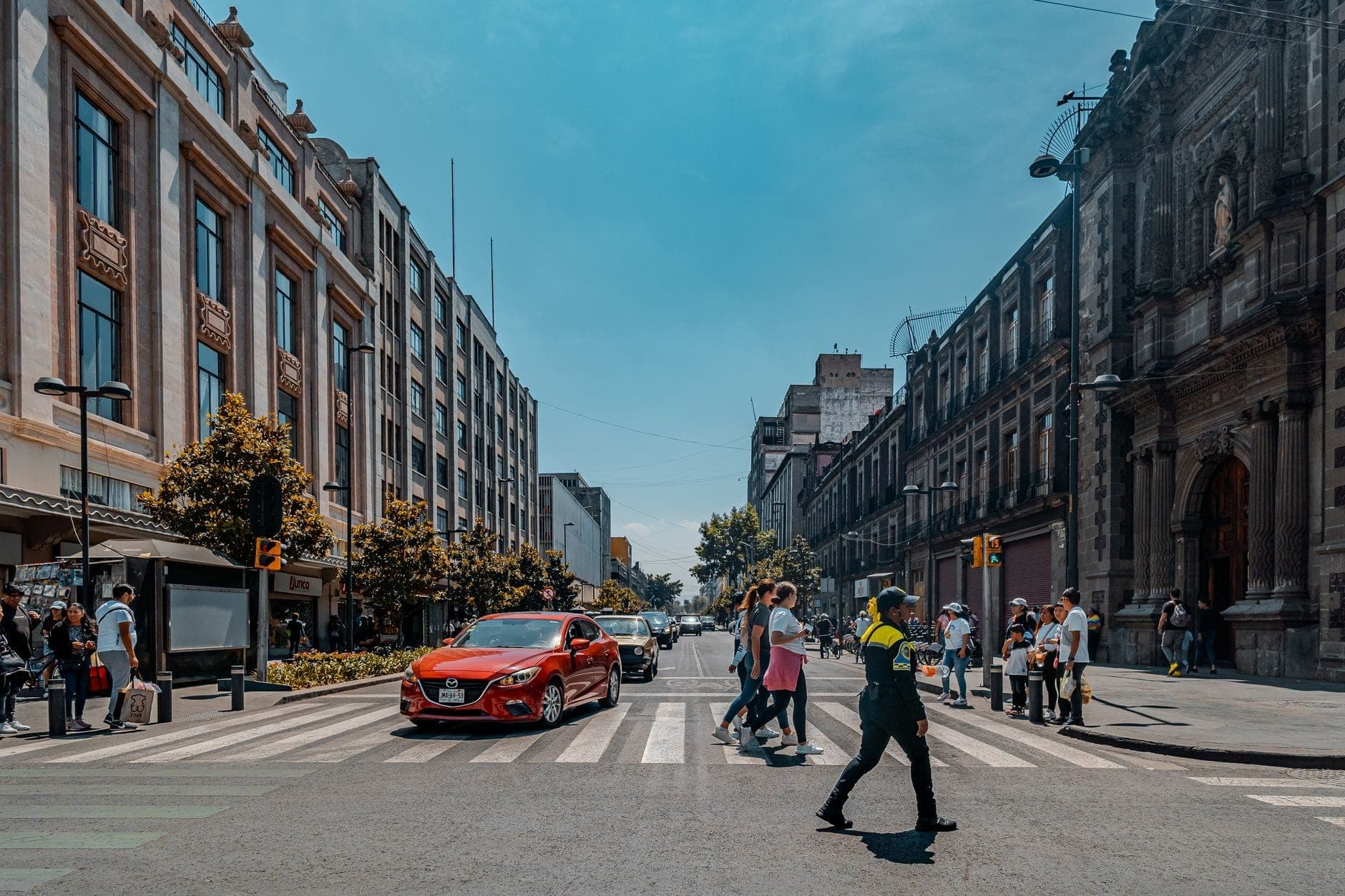 Busy crosswalk with pedestrians and a police officer in central Mexico City, surrounded by historic and modern buildings on a sunny day.