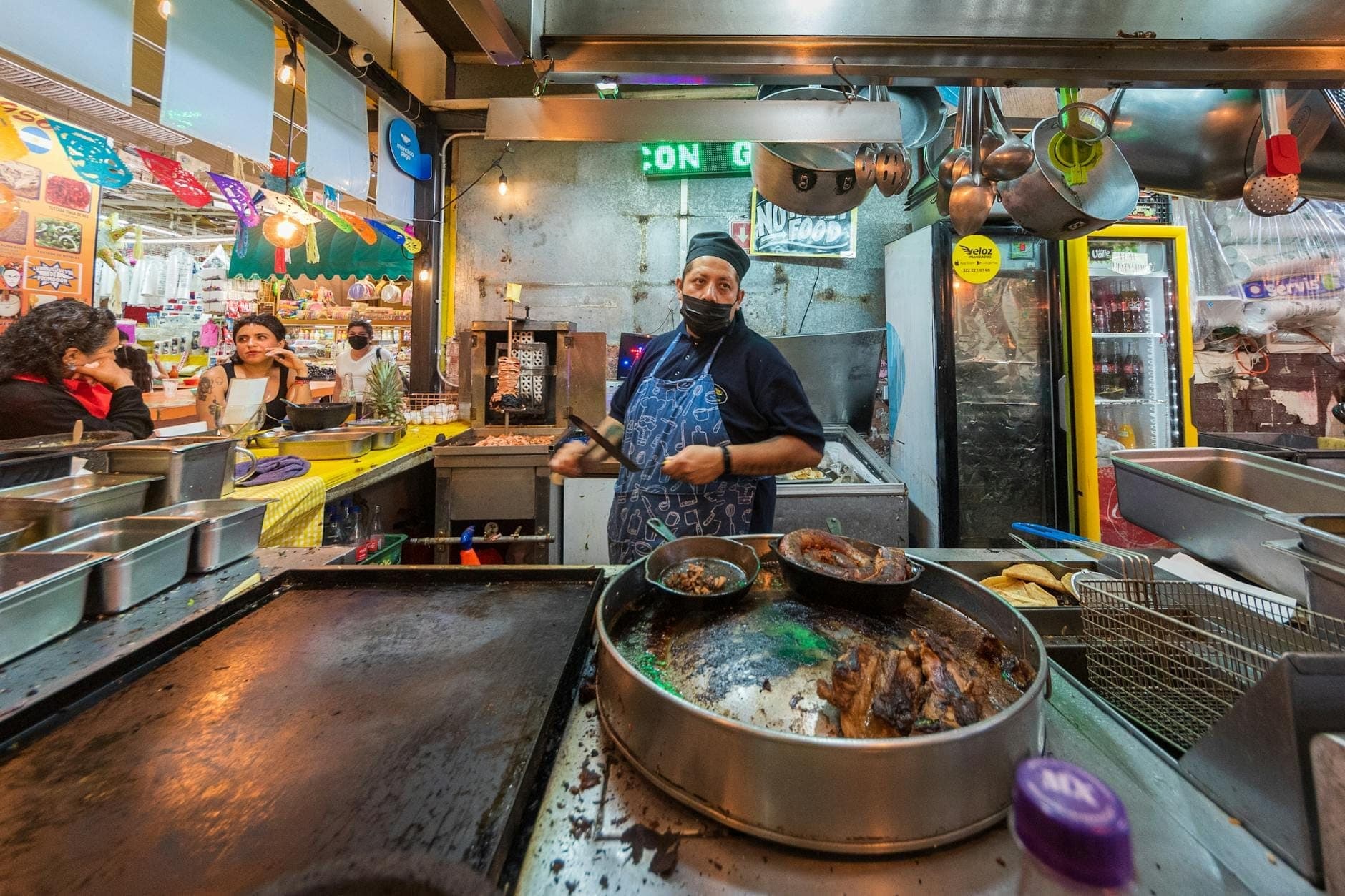 Un colorido y concurrido puesto de comida callejera en Ciudad de México con un vendedor cocinando detrás del mostrador, rodeado de varios ingredientes y utensilios de cocina.