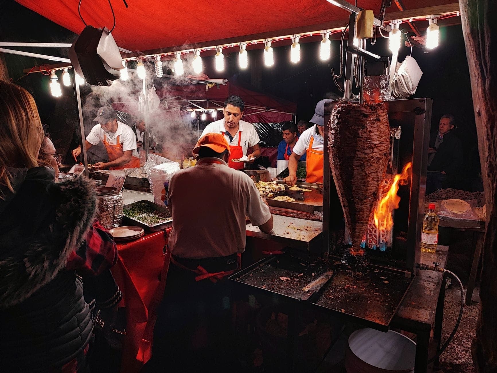 Imagen nocturna de un concurrido puesto de comida callejera en Ciudad de México con trabajadores preparando tacos al pastor, un trompo de carne y clientes esperando.