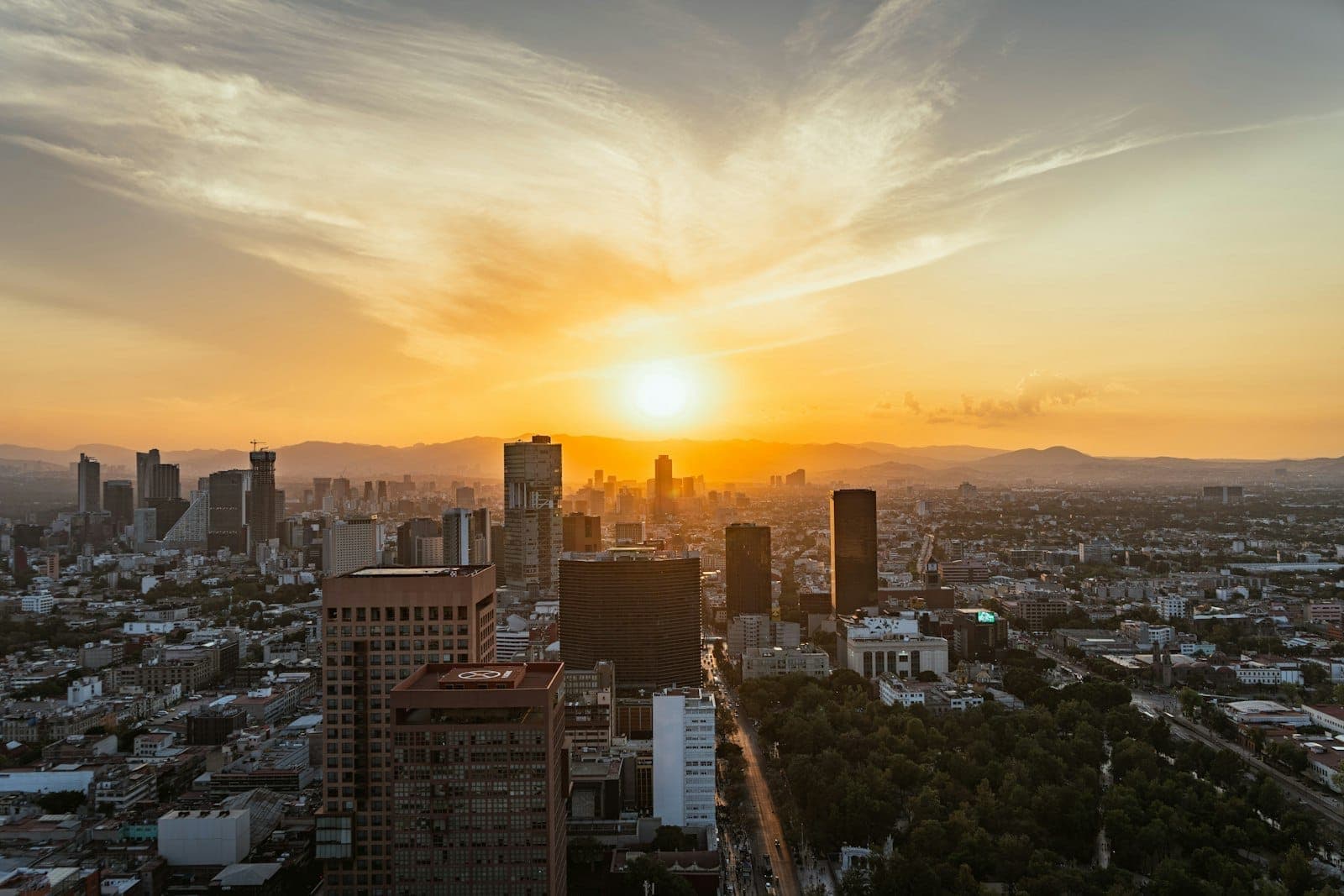 Stunning landscape view of Mexico City skyline at sunset with downtown skyscrapers, mountains in the background, and dramatic clouds above the city.