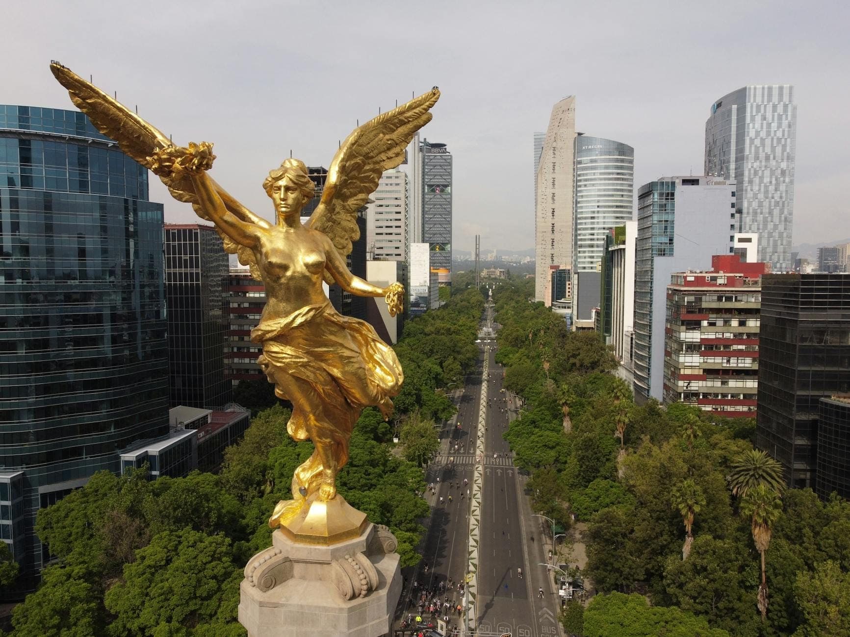 Widok z lotu ptaka na złotą statuę Ángel de la Independencia na szczycie kolumny, z obsadzoną drzewami aleją Paseo de la Reforma i nowoczesną panoramą miasta w tle.
