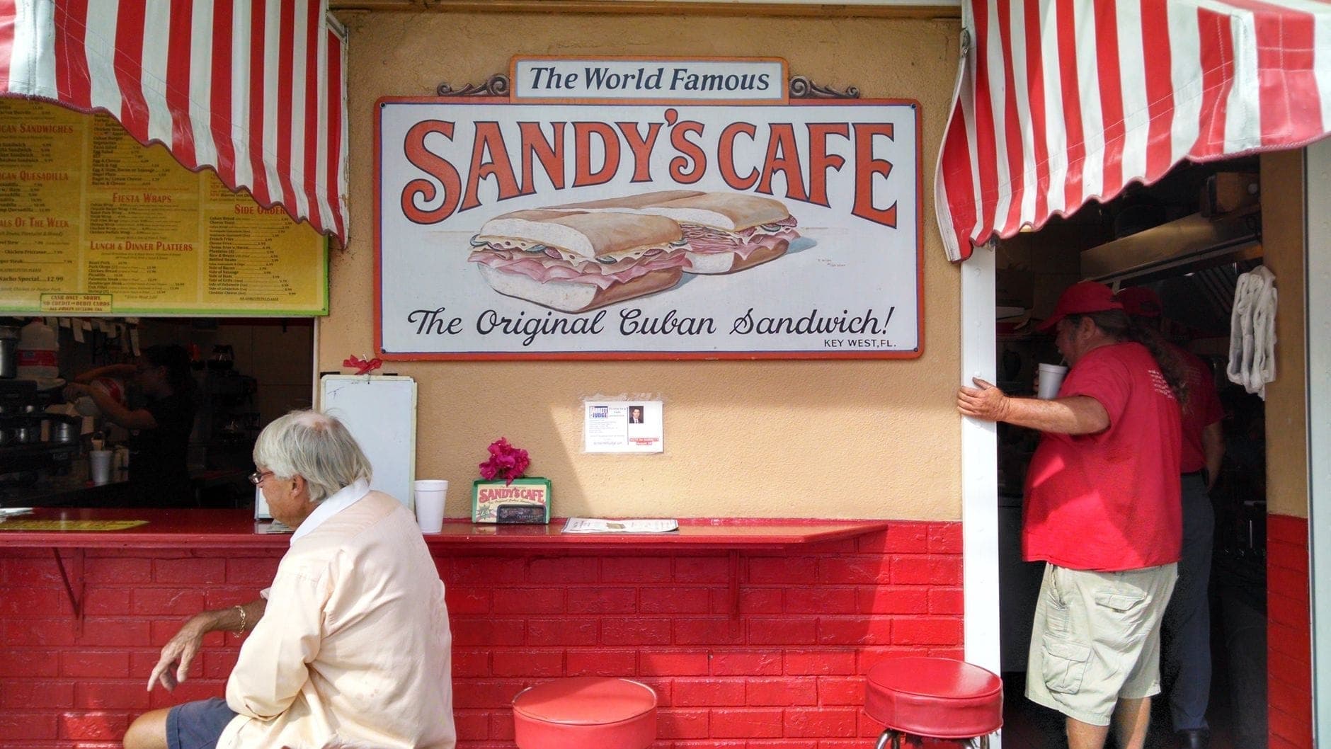 Streetfront counter of Sandy's Cafe with a sign advertising Cuban sandwiches and people sitting and ordering food.