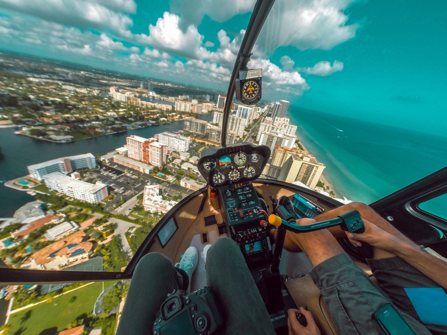A couple in a helicopter cockpit flying over Miami Beach, with the city, coastline, and turquoise ocean visible below.