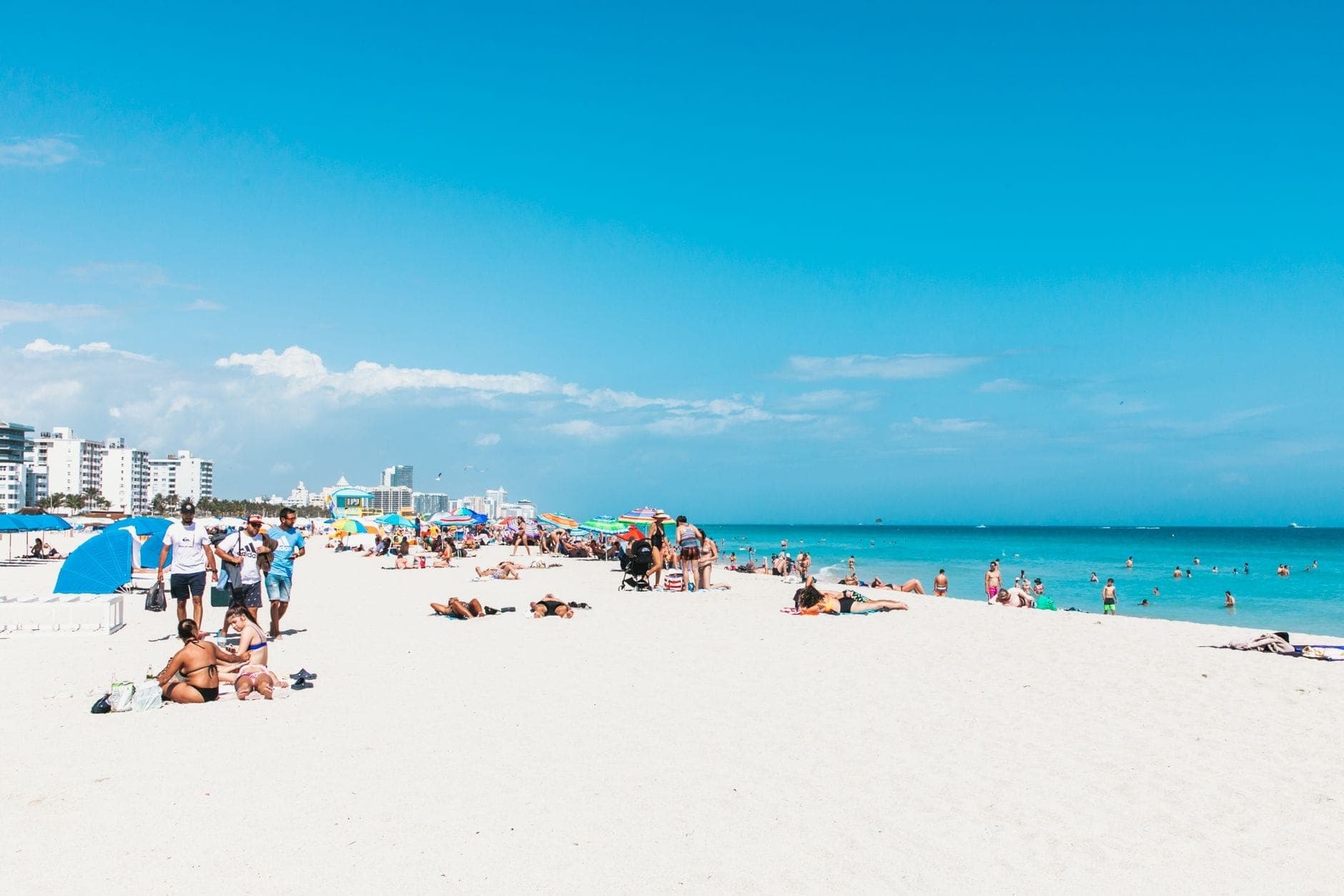 Crowded Miami beach scene with sunbathers, umbrellas, turquoise water, and city skyline under a clear blue summer sky.