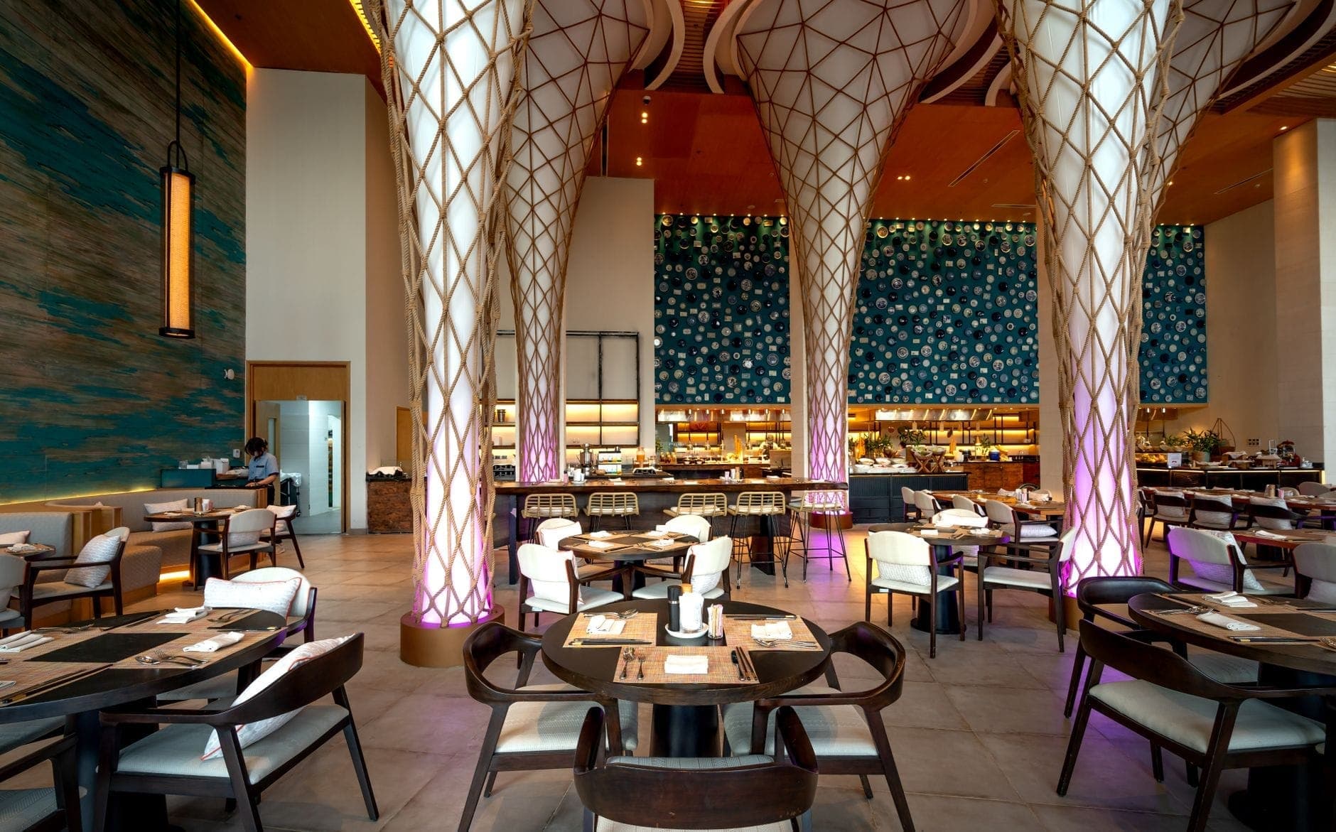 Intérieur spacieux et raffiné d'un restaurant avec une architecture moderne, d'élégantes colonnes et un éclairage stylisé, avec des tables bien dressées et un bar animé en arrière-plan.