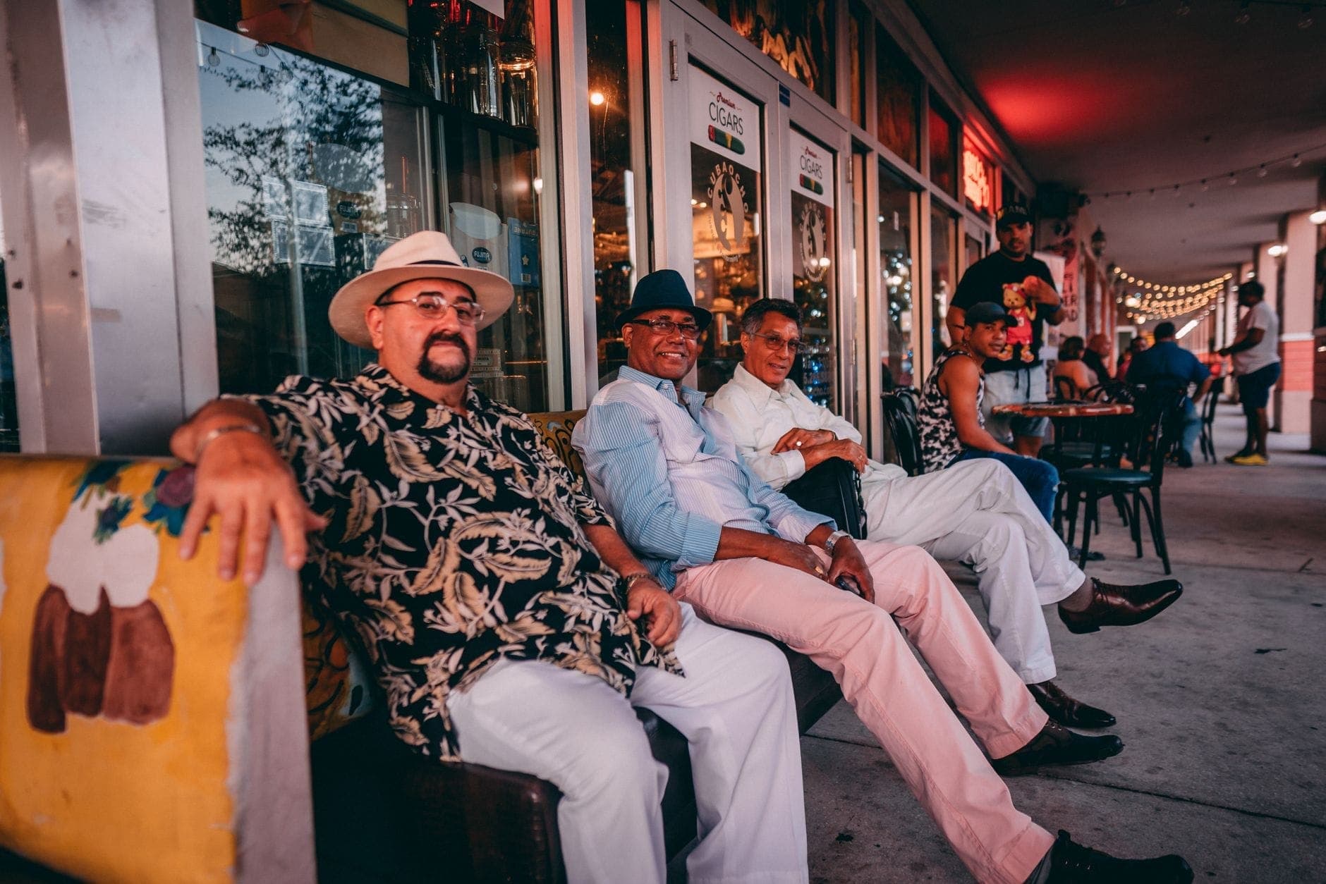 A group of people sitting outside a restaurant or café on a lively Miami street with festive evening lighting.
