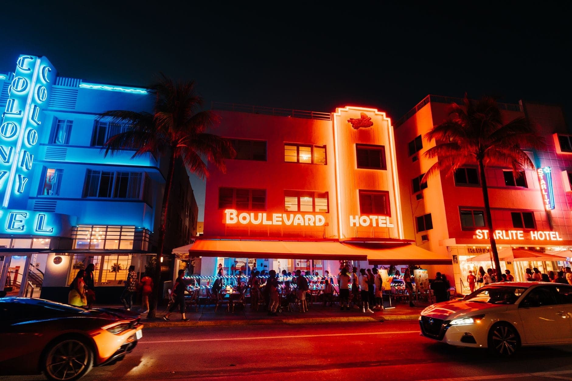 Night view of Miami's Ocean Drive with neon-lit hotels, crowds, and palm trees, illustrating a lively tourist area.