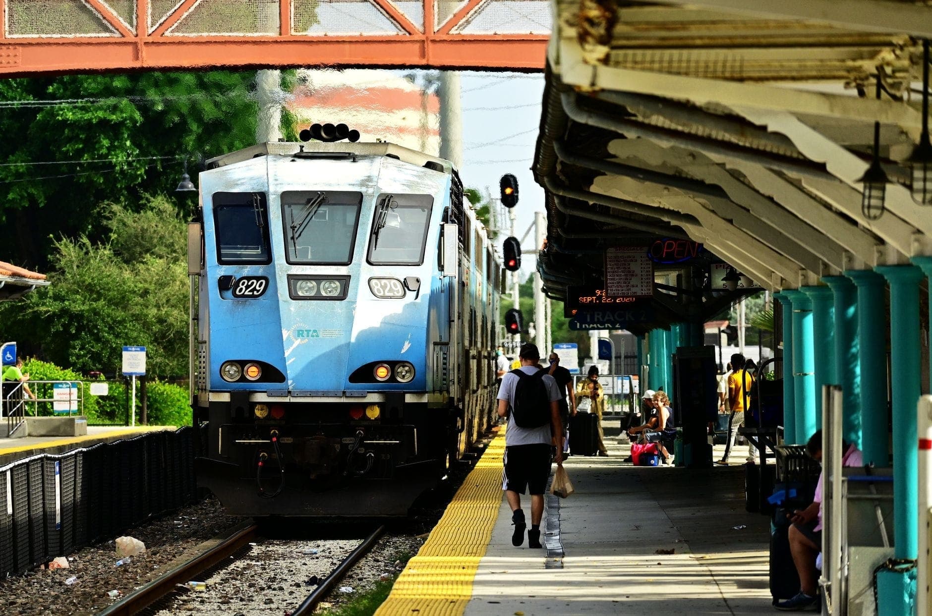 A commuter train arriving at a station with people walking on the platform, trees in the background, and a pedestrian overpass above.