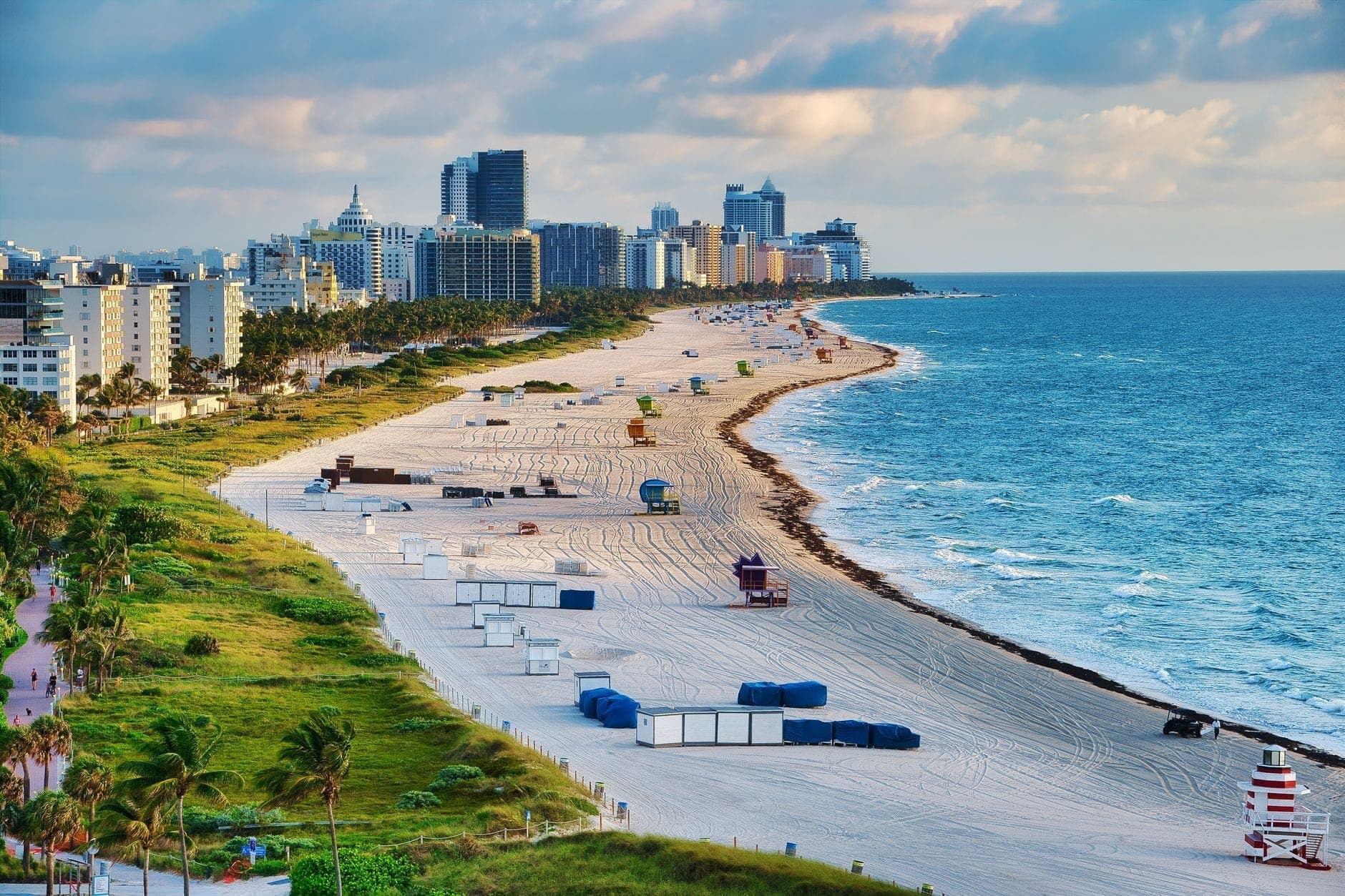 View of a sunny Miami Beach with a long stretch of sand, blue ocean, and city skyline in the background.