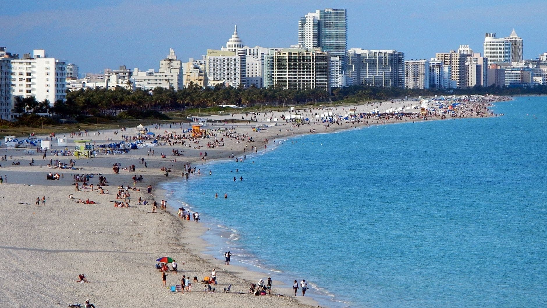 Wide view of Miami Beach with turquoise ocean, sandy shoreline, people swimming and sunbathing, and Miami skyline in the background.
