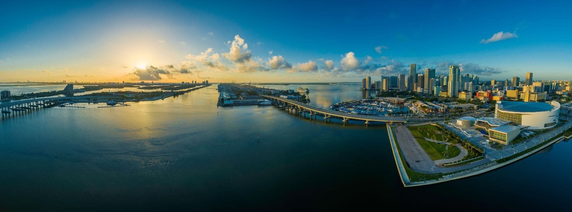 Wide panoramic aerial view of downtown Miami skyline, Biscayne Bay, and MacArthur Causeway at sunrise with clouds and water reflecting the sky.