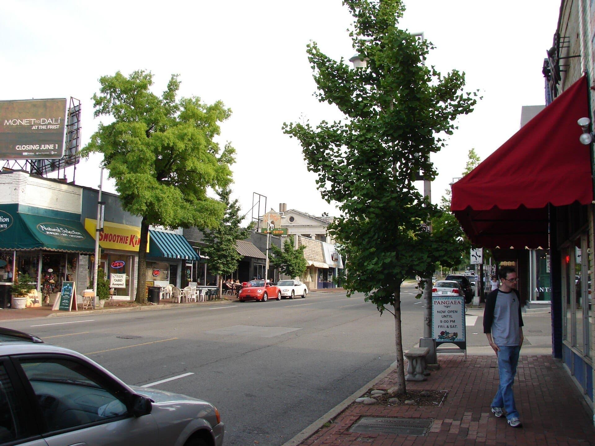 A street view of Hillsboro Village in Nashville showing local shops, a Smoothie King storefront, green trees, and a few parked cars on a sunny afternoon.