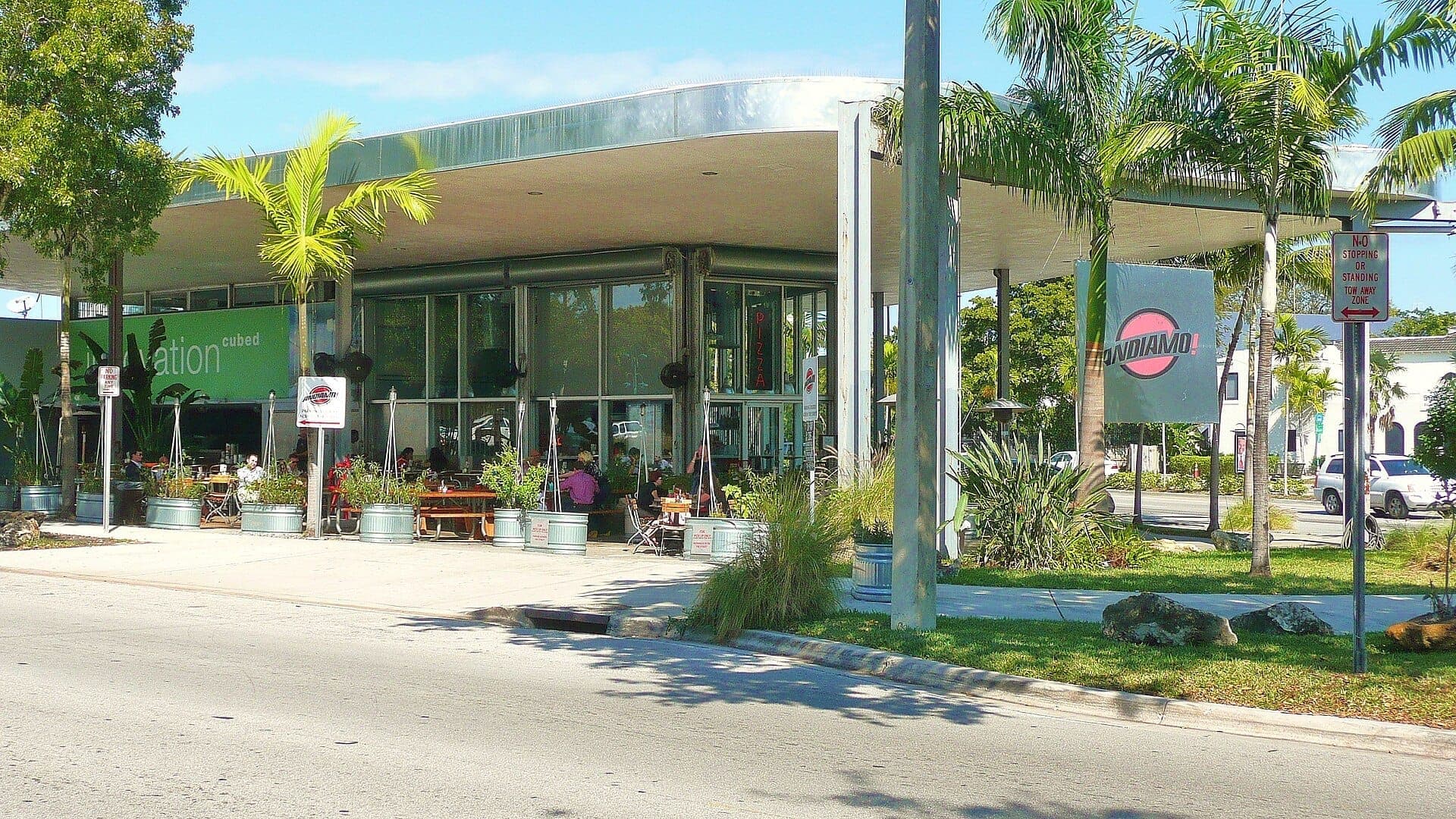 A modernist glass-walled building with a cantilevered roof and outdoor seating, located on Biscayne Boulevard in the MiMo Historic District.