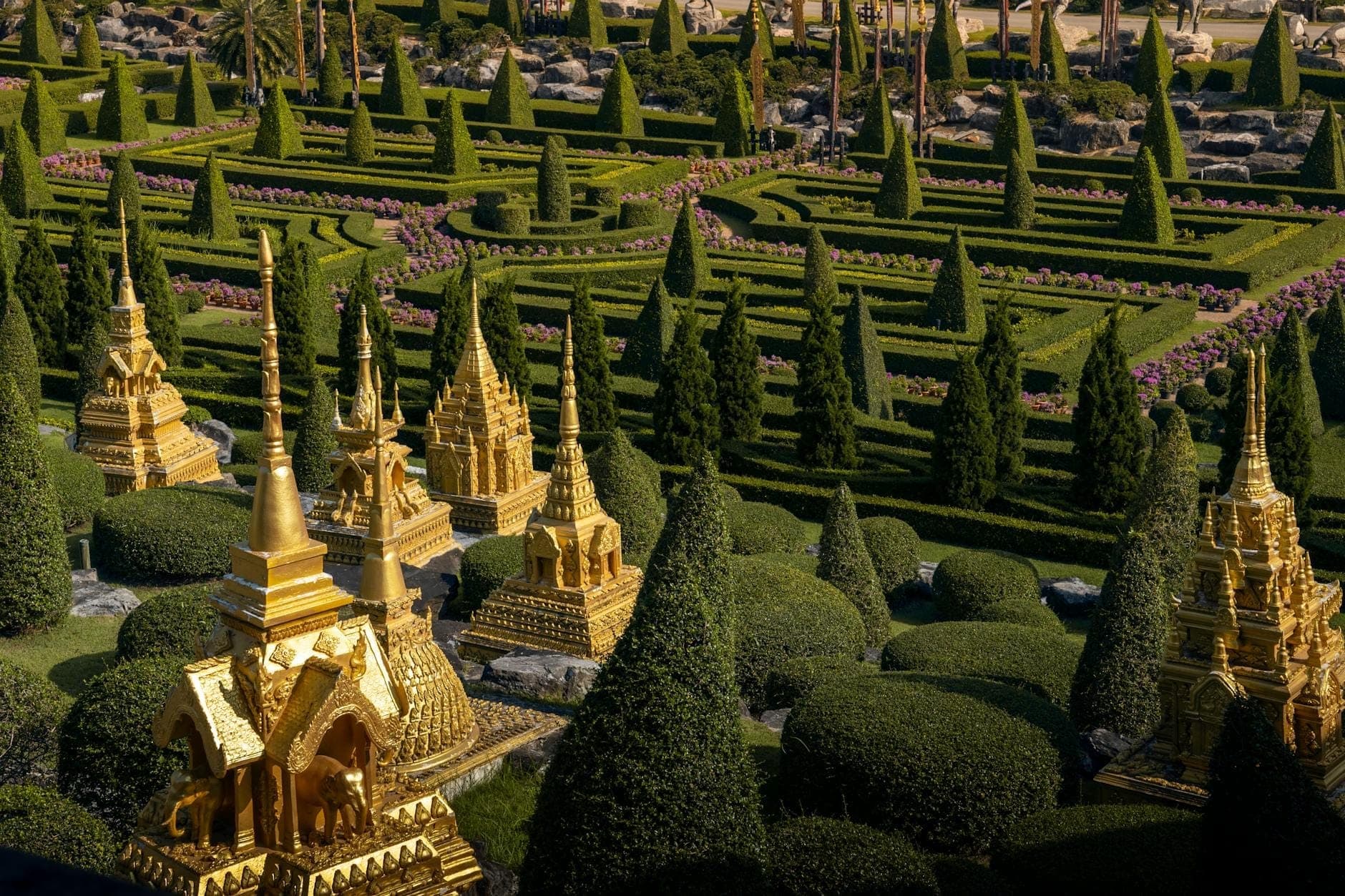 Miniature golden Thai temples arranged among manicured hedges and topiary trees, viewed from above in a sunlit open-air park setting.