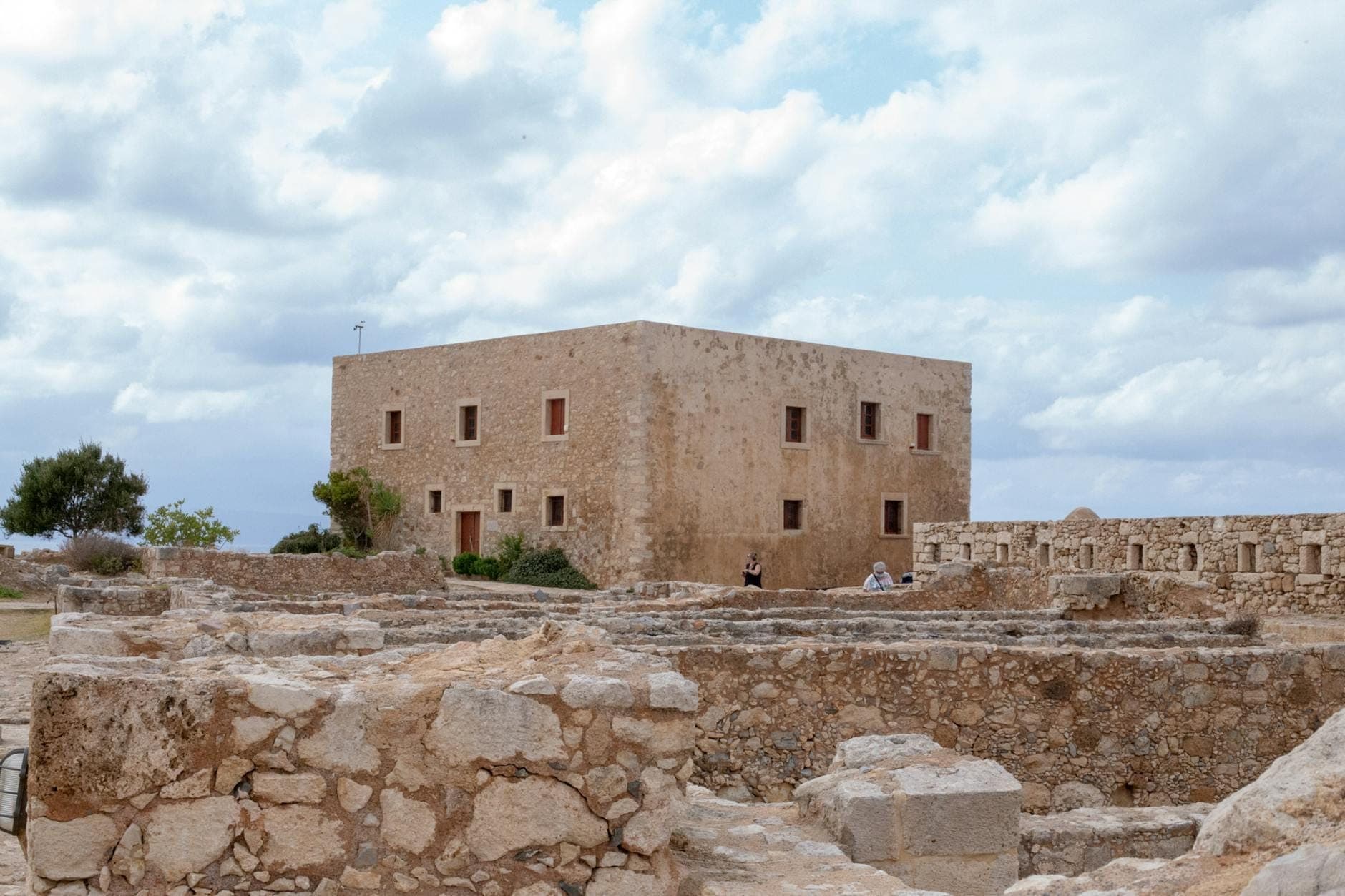 Alte Steinruinen mit einem großen rechteckigen venezianischen Gebäude im Hintergrund unter einem bewölkten Himmel auf Kreta.