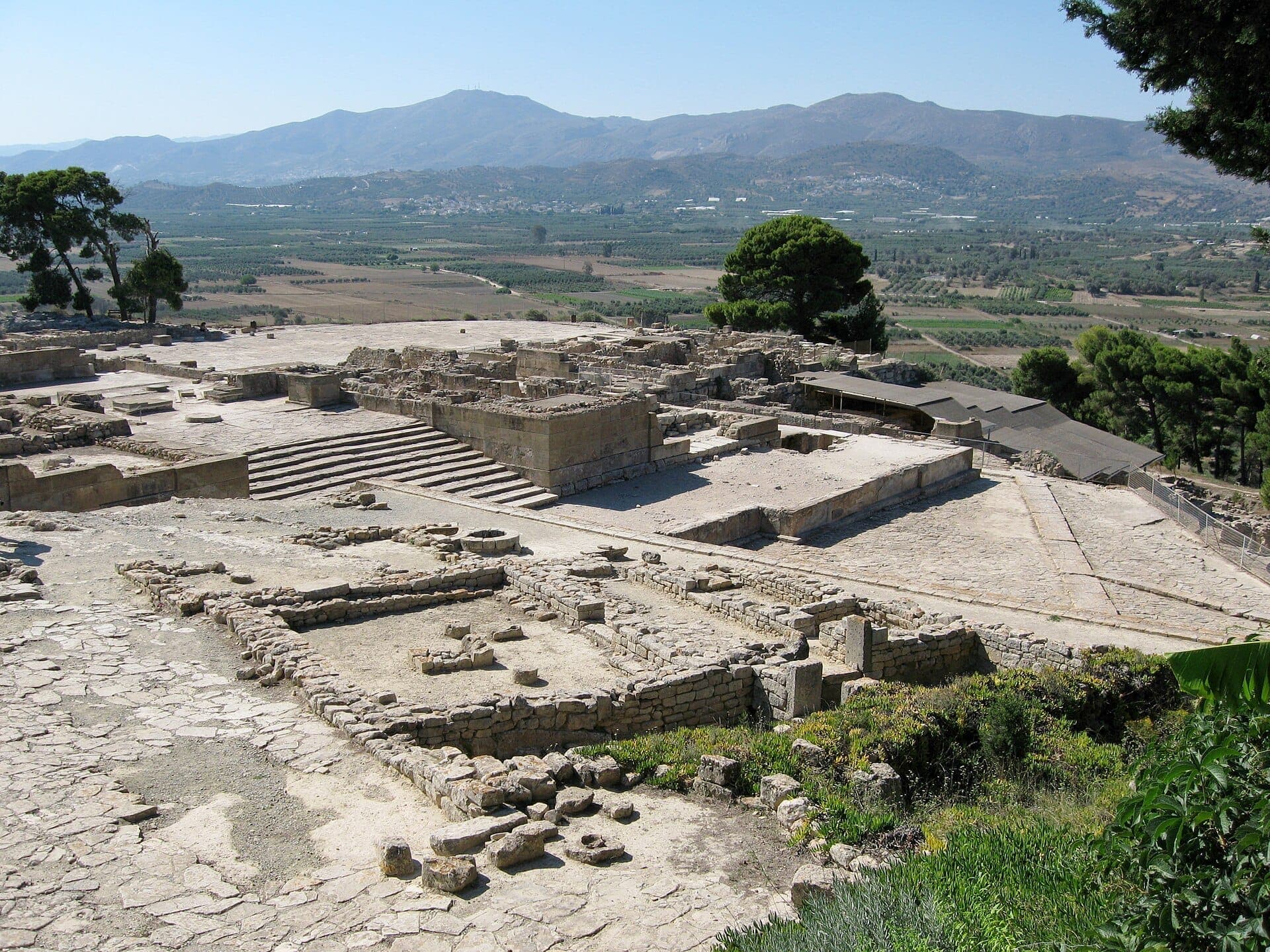 Die Ruinen des Palastes von Phaistos mit steinernen Fundamenten und Treppen überblicken die malerische Mesara-Ebene und die fernen Berge Kretas unter einem klaren Himmel.