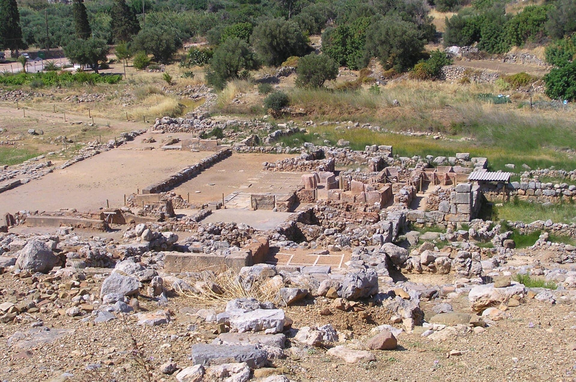 Blick auf die archäologischen Ruinen des Palastes von Zakros, umgeben von trockenem Gras, Felsen und grünen Bäumen bei hellem Tageslicht im östlichen Kreta.