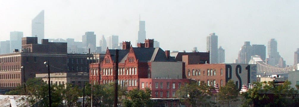 อาคารอิฐแดงทางประวัติศาสตร์ของ MoMA PS1 ตั้งอยู่ท่ามกลางโครงสร้างอุตสาหกรรมใน Queens โดยมีเส้นขอบฟ้าของ New York City ปรากฏให้เห็นในพื้นหลัง