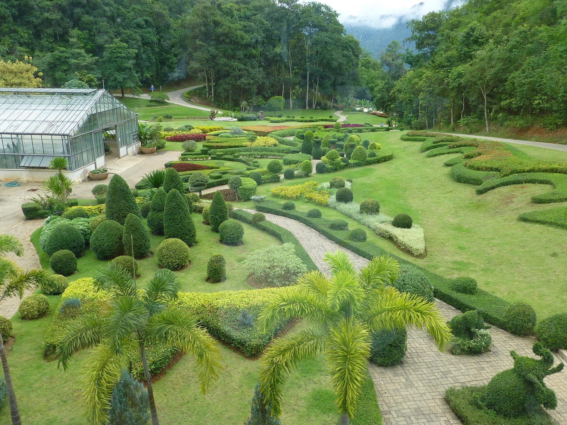 Giardini curati e una serra di vetro a Mon Cham, Chiang Mai, circondati da colline lussureggianti e sinuosi sentieri di pietra sotto un cielo nuvoloso.