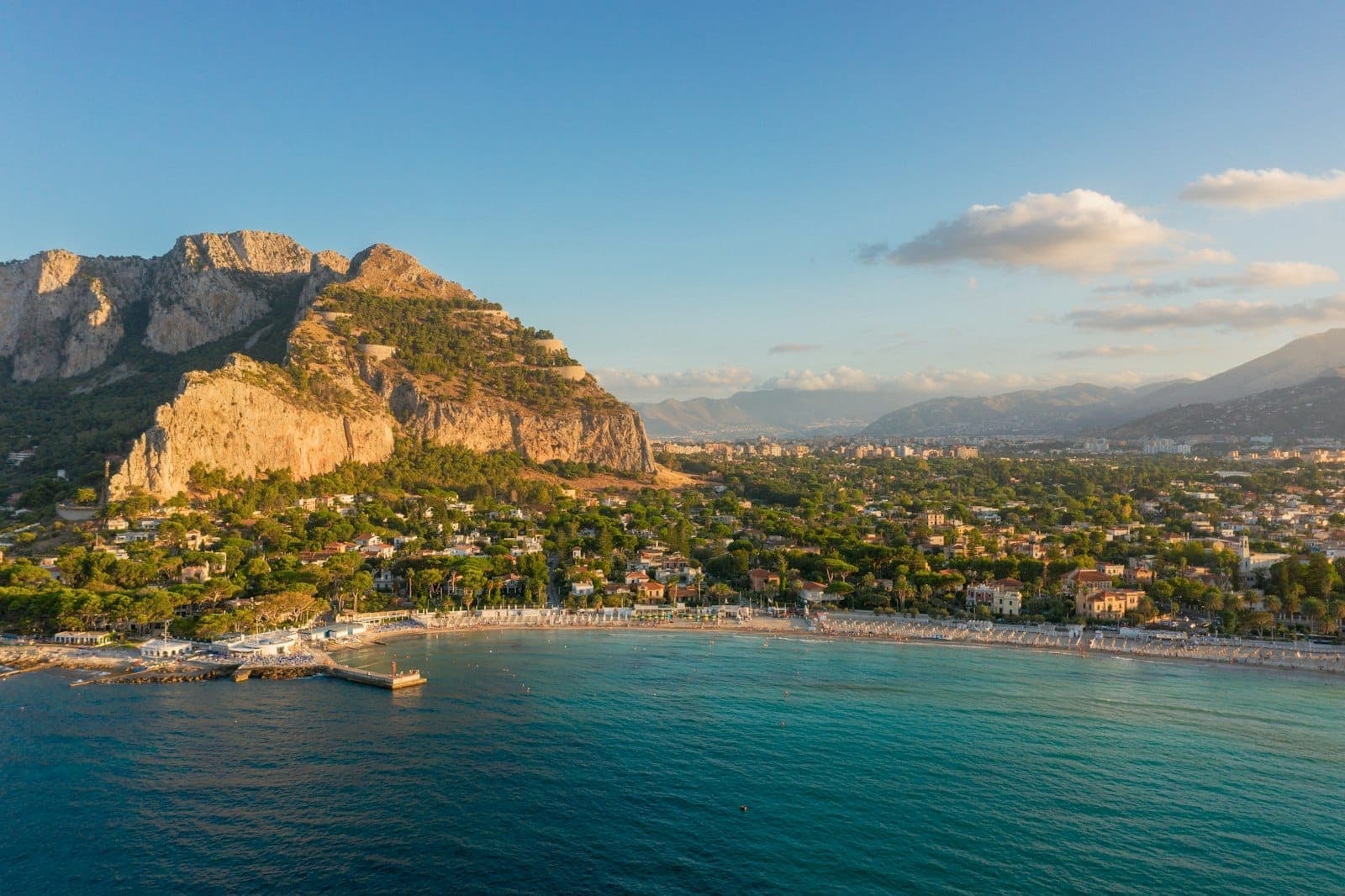 Pemandangan udara Pantai Mondello dengan pasir putih, air biru toska, dan pepohonan hijau rimbun, diapit Monte Pellegrino saat matahari terbenam di dekat Palermo.
