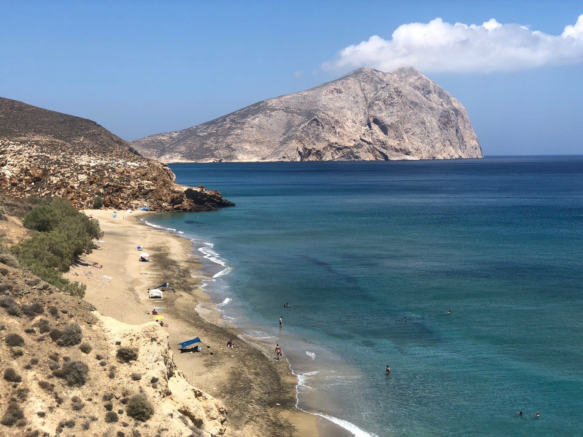 Der Strand Monolithos mit schwarzem Sandufer, einigen Sonnenbadenden, sanften Wellen, blauem Wasser, felsigen Klippen und einem großen Berg im Hintergrund.
