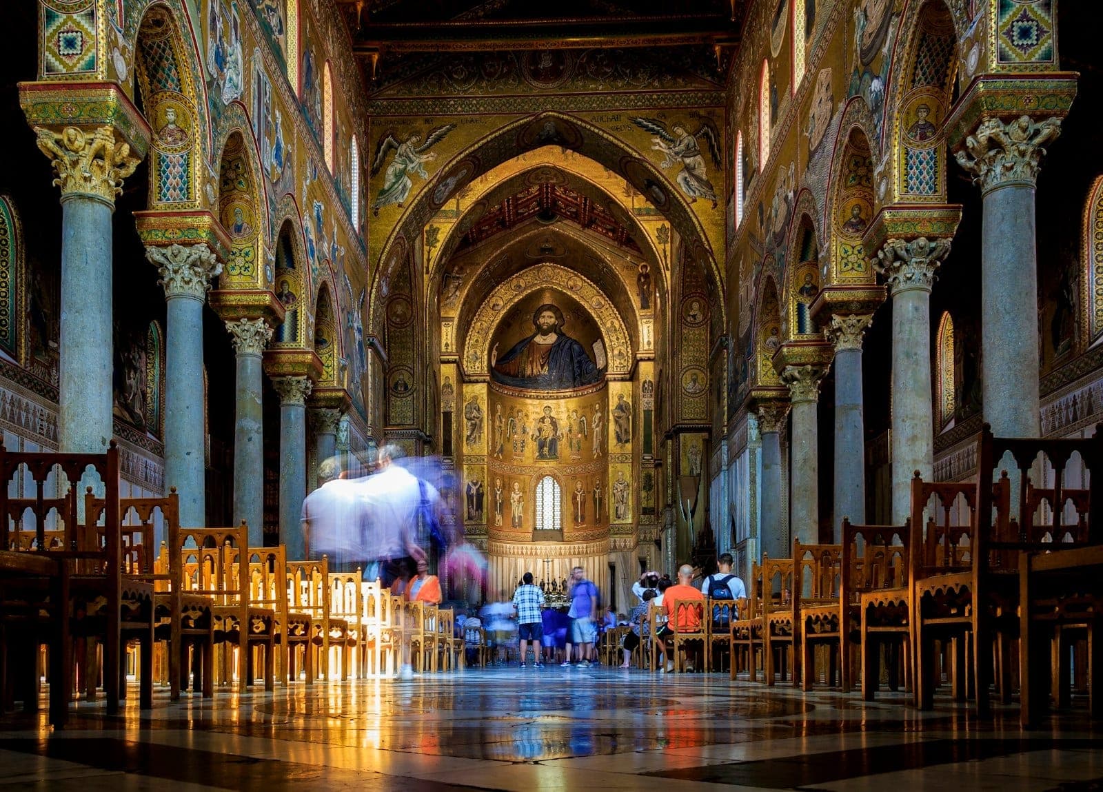 Pemandangan sepanjang nave Katedral Monreale dengan pengunjung dan apse yang ditutupi mosaik Bizantium emas berkilauan di latar belakang.