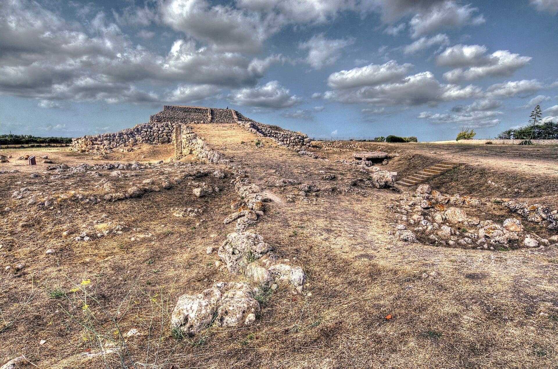 Rampa de piedra y estructura de altar de Monte d'Accoddi bajo un cielo dramático, rodeada de hierba seca y rocas dispersas en Cerdeña.