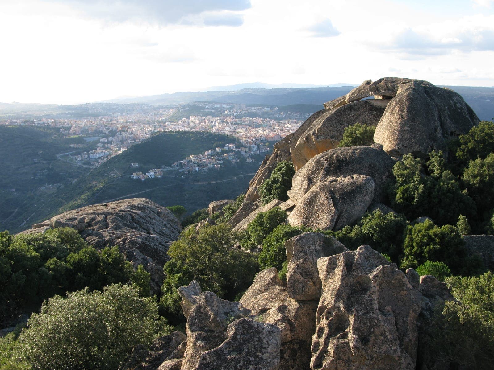 Vista desde el Monte Ortobene con rocas de granito escarpadas, bosque verde y la ciudad de Nuoro extendiéndose en el lejano valle de abajo.