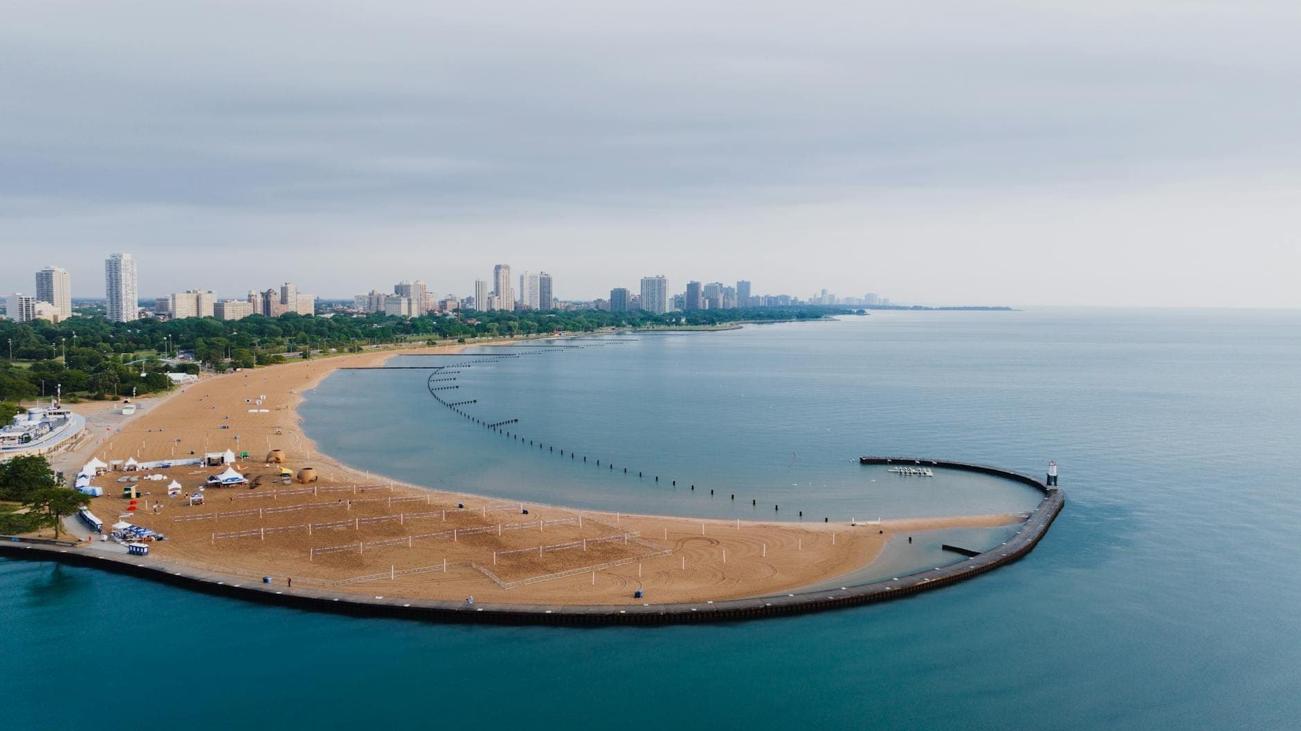 A wide sandy beach with volleyball courts, clear calm water, and Chicago’s skyline in the distance under a cloudy sky.