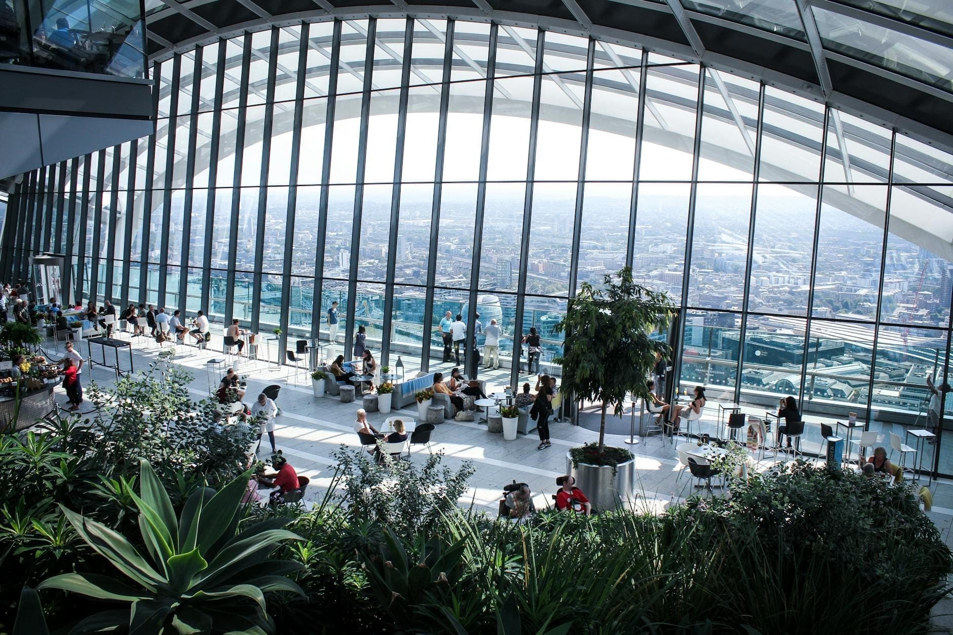 ภายใน Sky Garden ในลอนดอน พร้อมหน้าต่างกระจกขนาดใหญ่ พืชพรรณเขียวขจี และนักท่องเที่ยวที่เพลิดเพลินกับวิวพาโนรามาของเส้นขอบฟ้าเมือง
