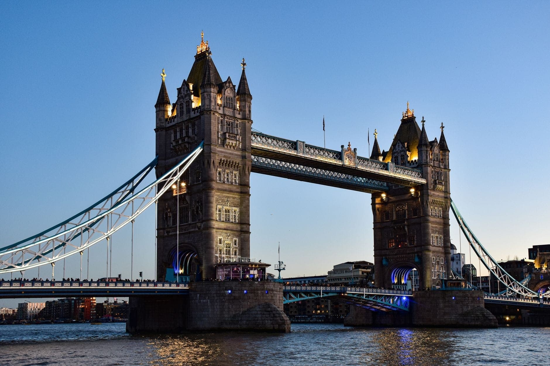 Tower Bridge ในลอนดอนยามพลบค่ำ สว่างไสวเต็มที่ มีท้องฟ้าสีฟ้าและแม่น้ำเทมส์อยู่เบื้องหน้า สะท้อนให้เห็นสัญลักษณ์ที่โดดเด่นที่สุดของเมือง
