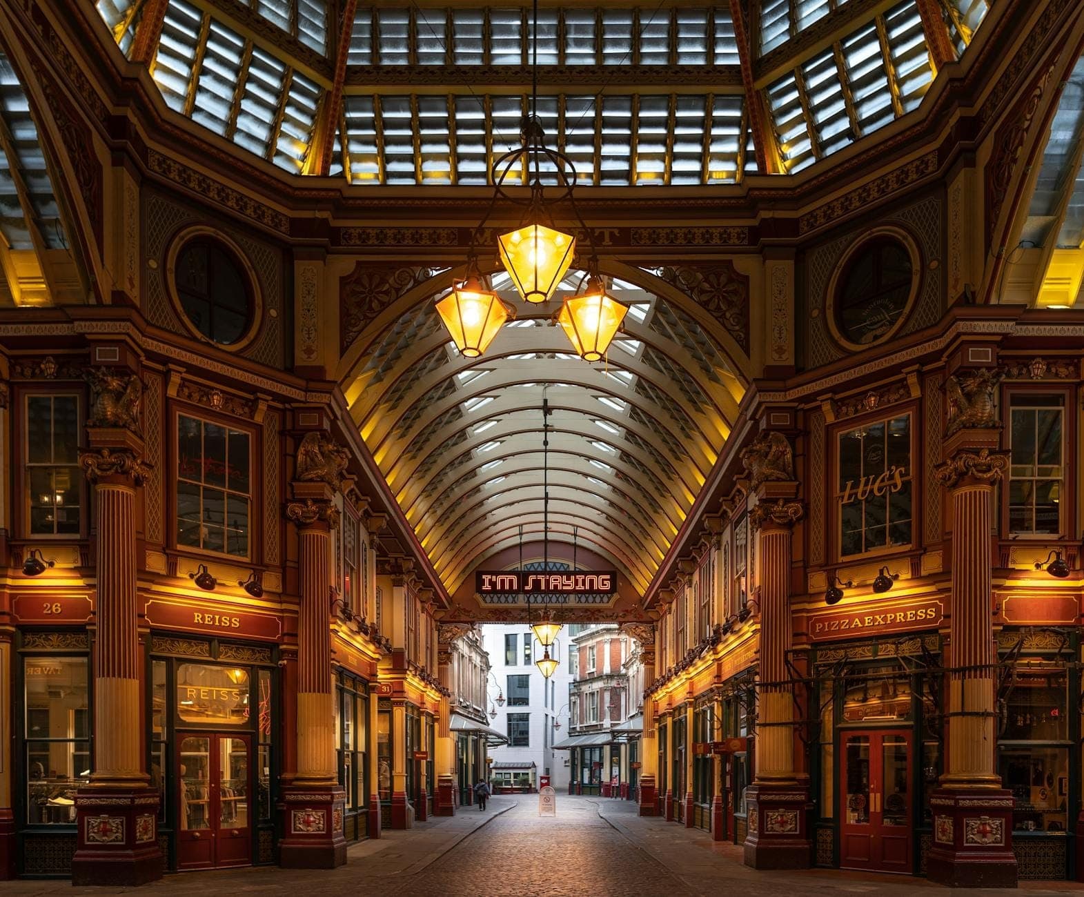 มุมมองแบบไวด์แองเกิลของภายในตลาด Leadenhall Market ในลอนดอนที่มีหลังคากระจกอันวิจิตรงดงาม พร้อมเสาสไตล์วิกตอเรียนที่โดดเด่น โคมไฟแขวน และซุ้มโค้งที่ตกแต่งอย่างหรูหรา