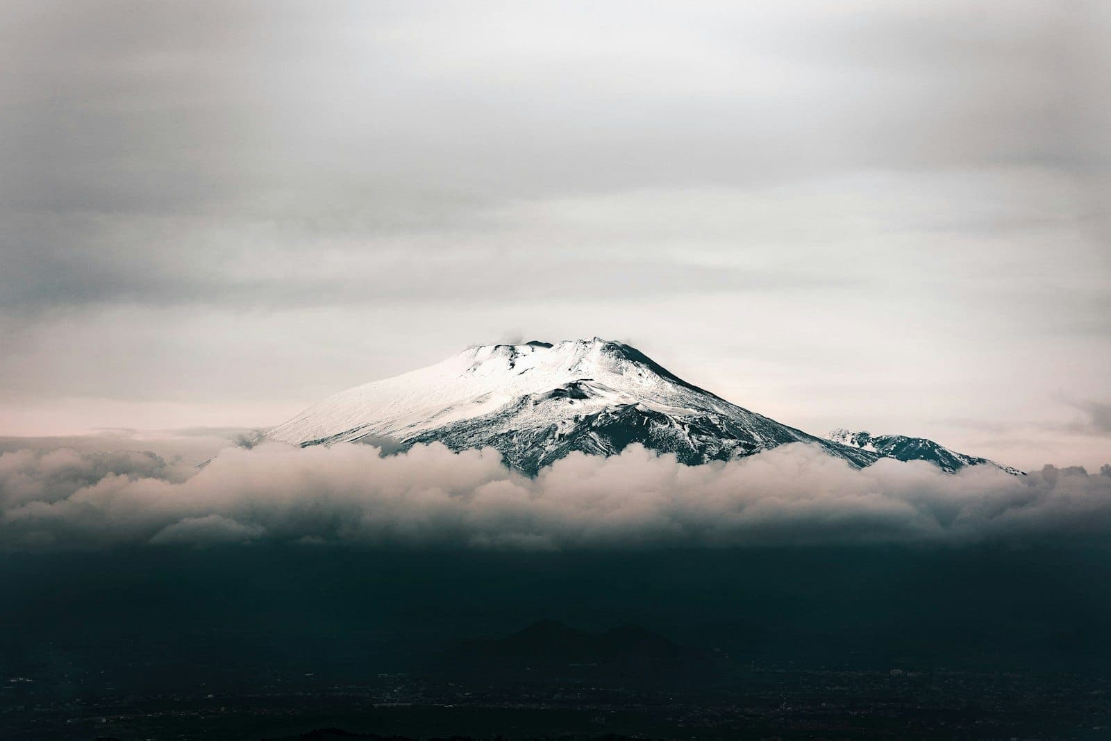 Puncak Gunung Etna yang bersalju menjulang di atas awan dengan langit yang dramatis dan lereng gunung berapi yang terlihat jelas, menjadi penanda alam yang mencolok di lanskap Sisilia.
