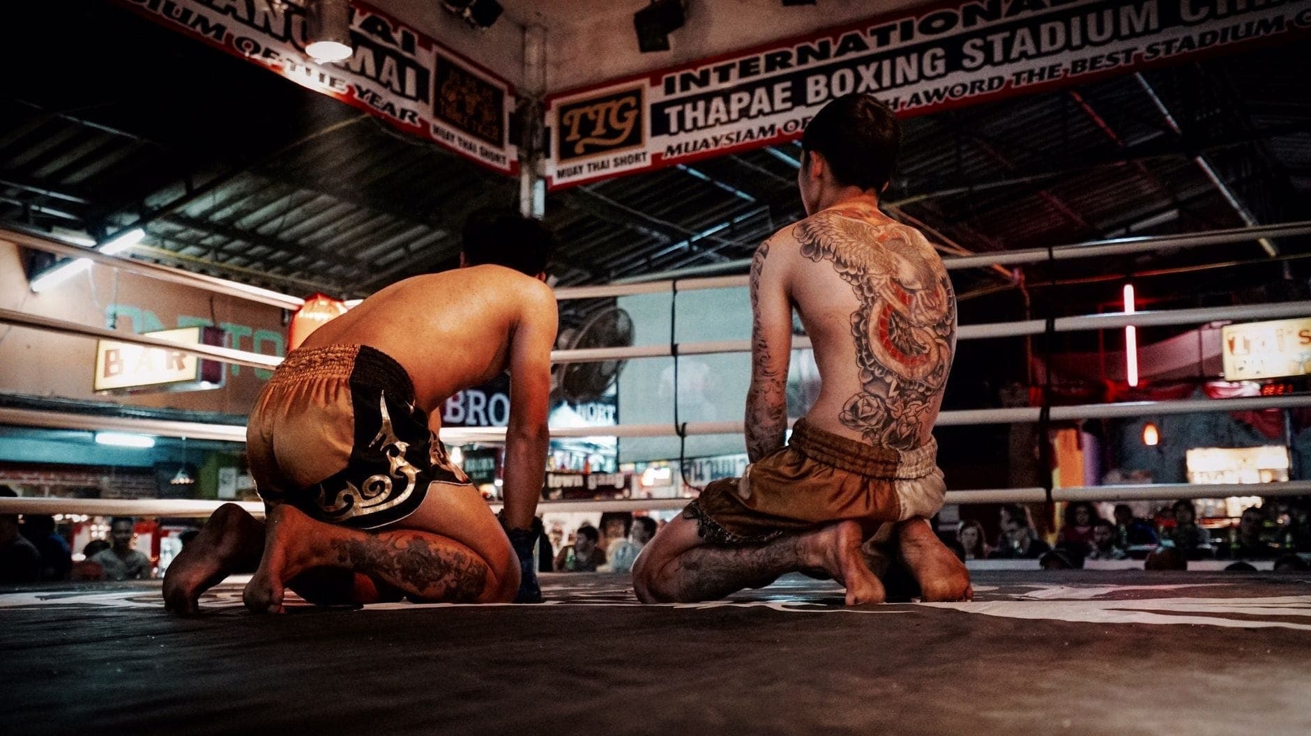 Dwóch zawodników Muay Thai klęczy na ringu w Thapae Boxing Stadium w Chiang Mai, otoczonych przez publiczność i oznakowanie obiektu.