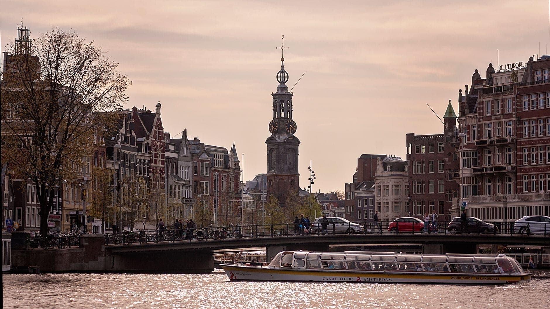 View of the Munttoren (Mint Tower) in Amsterdam, rising above canal houses and a busy bridge with a tour boat on the Amstel River at sunset.