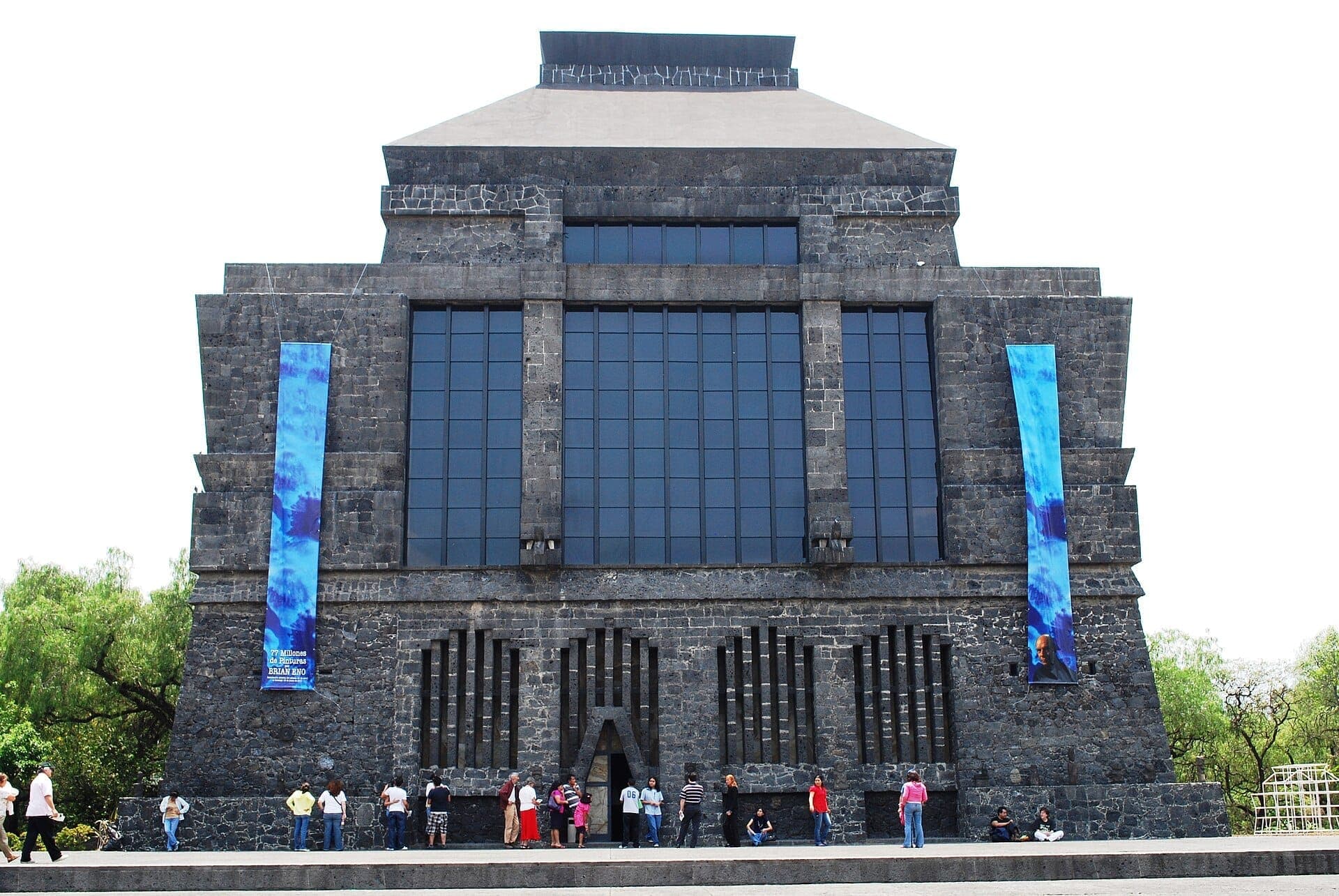 มุมมองด้านหน้าของ Museo Anahuacalli ที่แสดงให้เห็นผนังหินภูเขาไฟ หน้าต่างขนาดใหญ่ ป้ายแบนเนอร์สีน้ำเงิน และนักท่องเที่ยวที่ยืนอยู่ด้านนอกในวันที่แดดจัด
