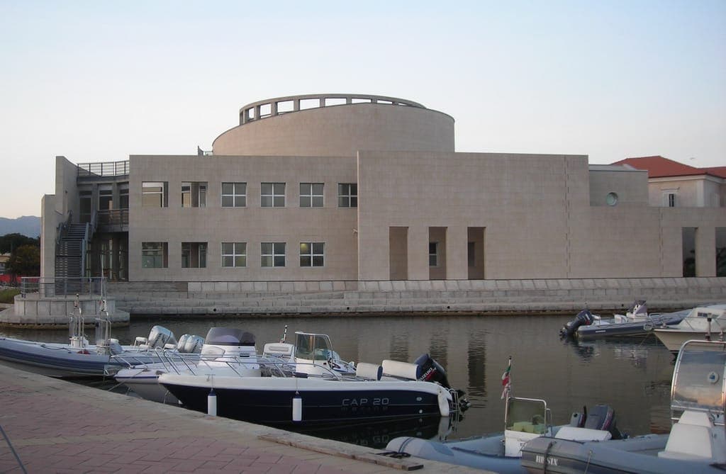 Vista frontal del Museo Archeologico di Olbia, un moderno edificio de museo de color beige con líneas geométricas, reflejado en las tranquilas aguas del puerto antiguo.