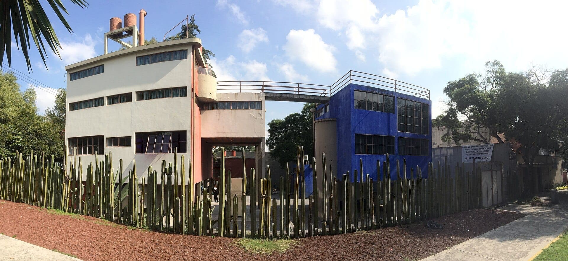 Museo Casa Estudio Diego Rivera y Frida Kahlo พร้อมอาคารโมเดิร์นนิสต์สีแดง ขาว และน้ำเงินที่เชื่อมต่อกัน และรั้วต้นกระบองเพชรสูงใต้ท้องฟ้าแดดจ้า