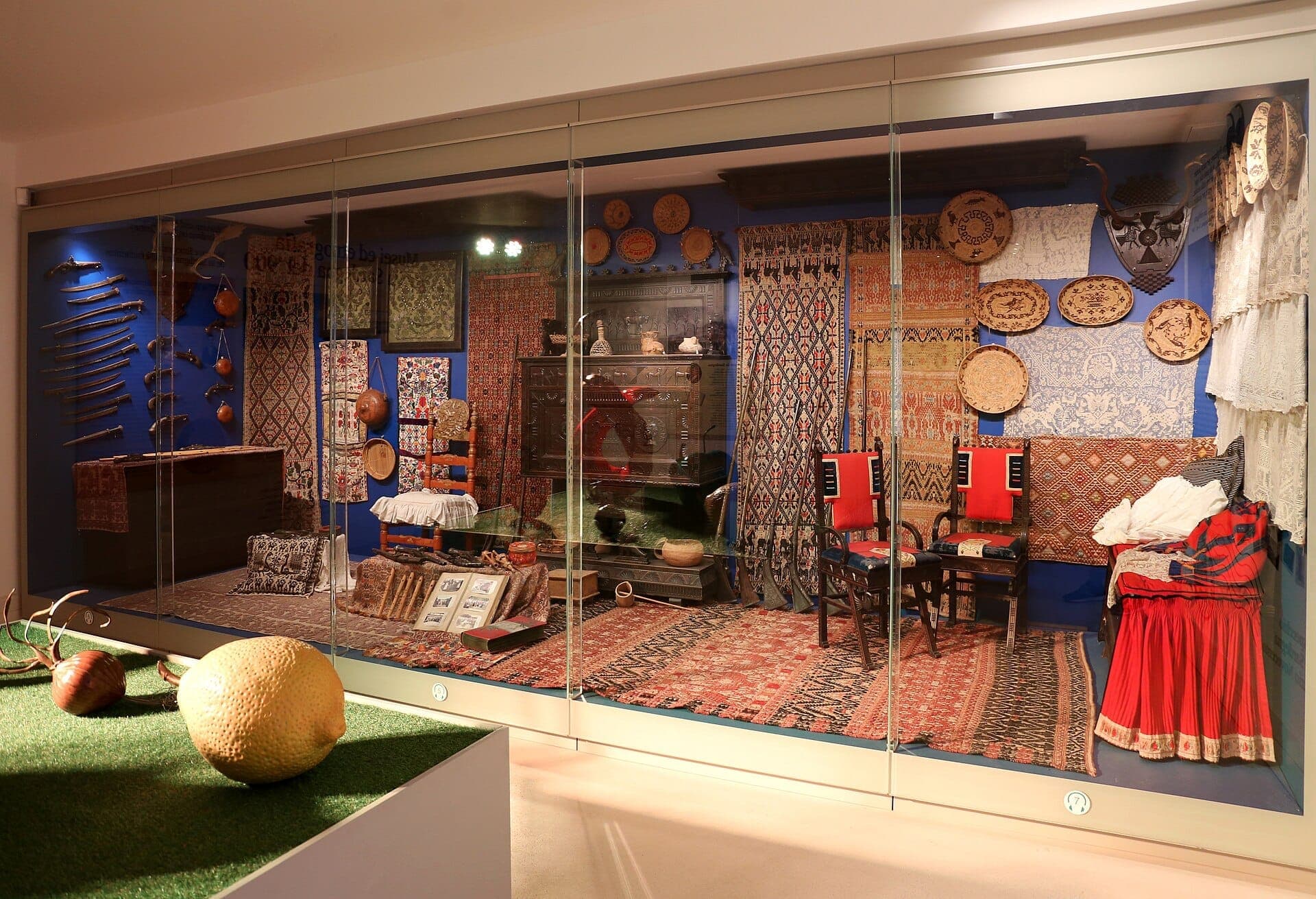 Una exposición muy iluminada en el Museo del Costume Nuoro muestra textiles sardos tradicionales, trajes populares, muebles de madera tallada y cestas tejidas detrás de un cristal.