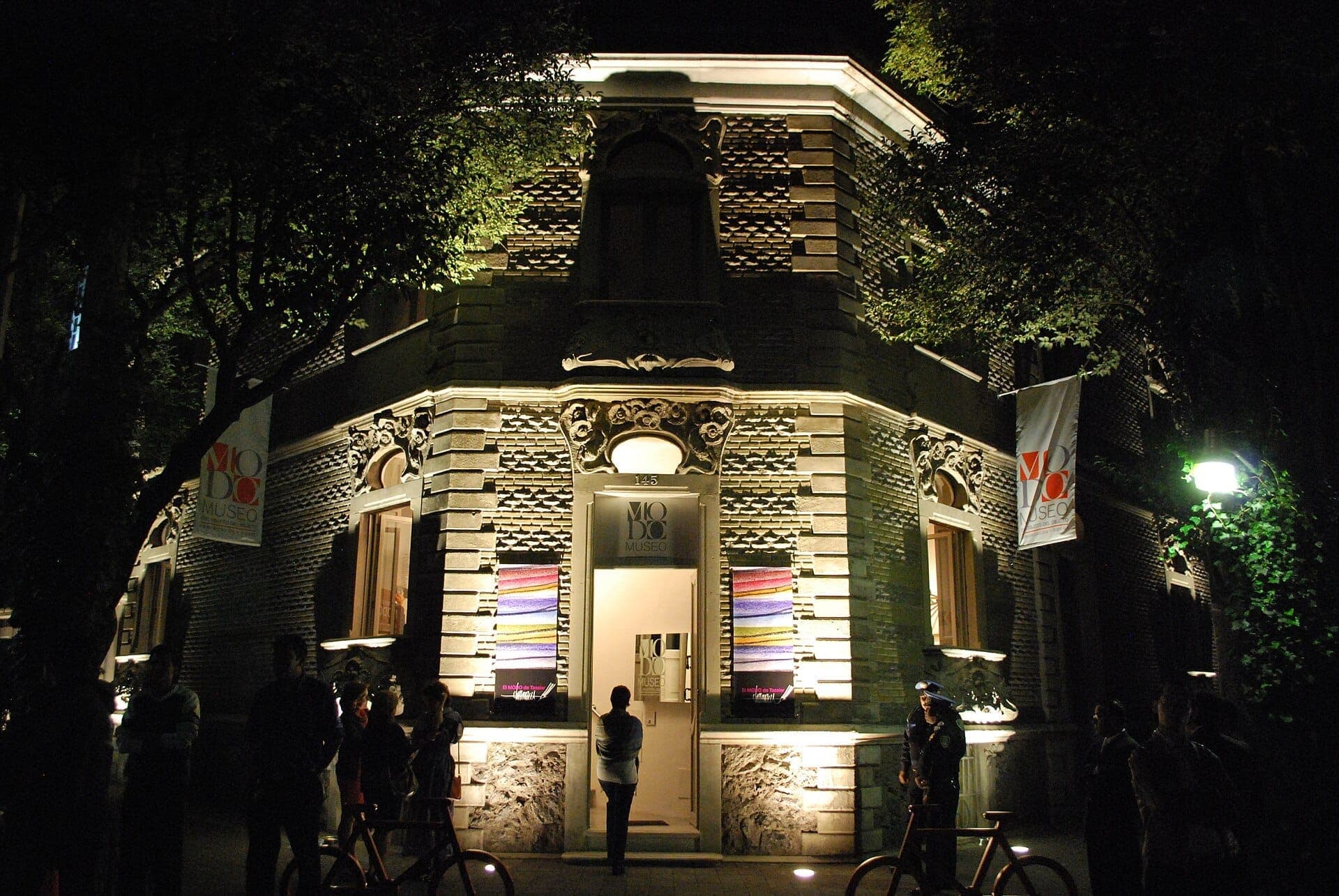 ด้านหน้าอาคารที่สว่างไสวของ Museo del Objeto del Objeto ในเม็กซิโกซิตี้ พร้อมรายละเอียดสไตล์ Art Nouveau และผู้คนที่มารวมตัวกันด้านนอกในเวลากลางคืน