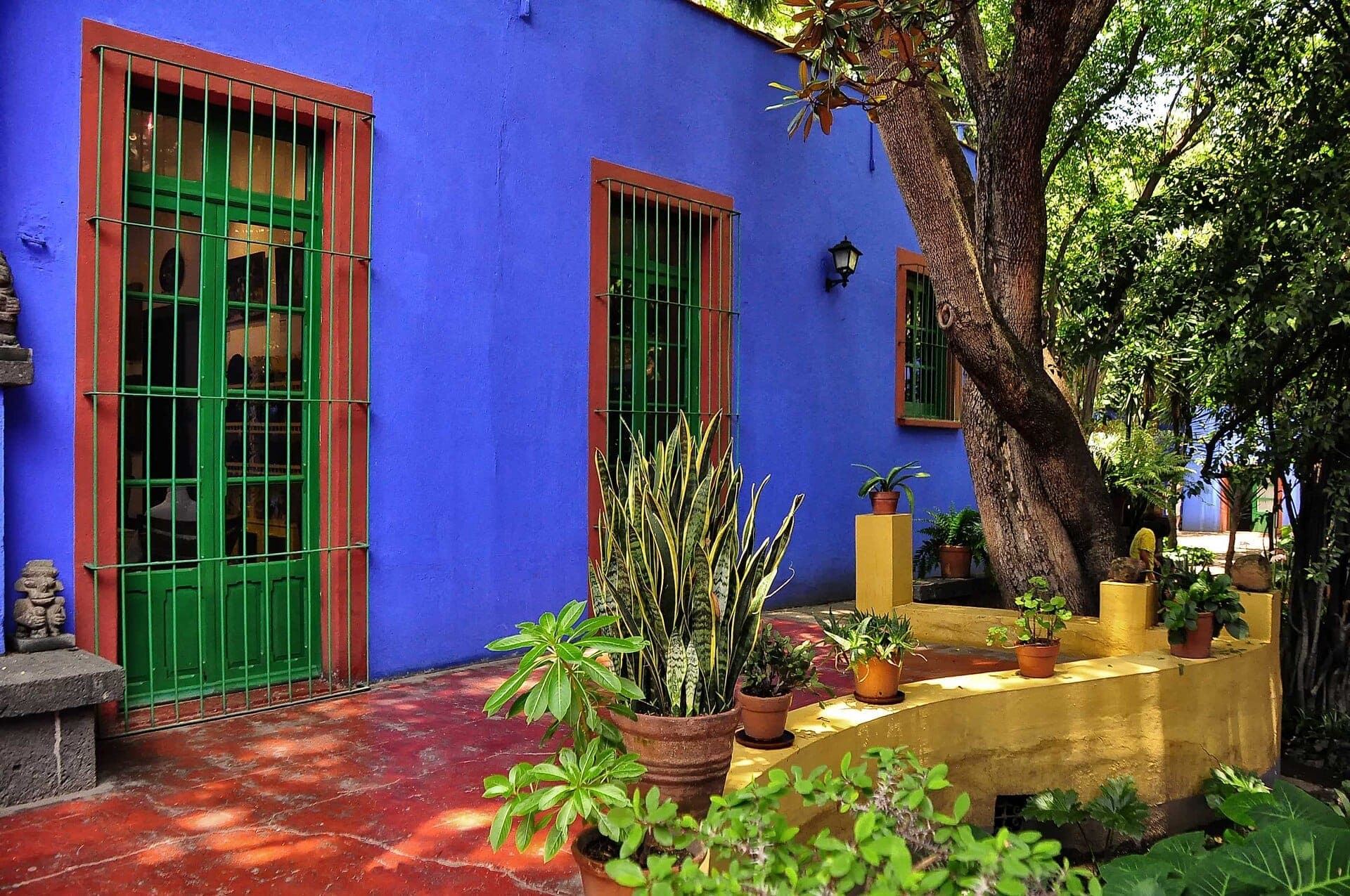 ด้านหน้าอาคารสีน้ำเงินสดใสของ Museo Frida Kahlo พร้อมหน้าต่างลูกกรงสีเขียว ต้นไม้ในกระถาง ต้นไม้ให้ร่มเงา และแสงแดดส่องผ่านในลานบ้าน
