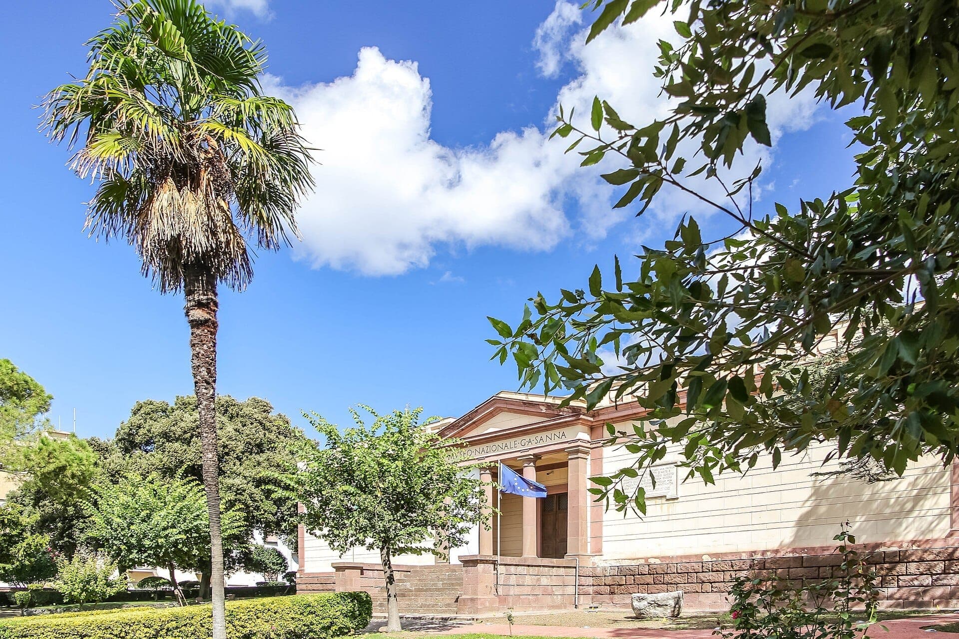 El Museo Nazionale G.A. Sanna en Sassari, Cerdeña, se ve detrás de exuberantes jardines, una alta palmera y bajo un brillante cielo azul.