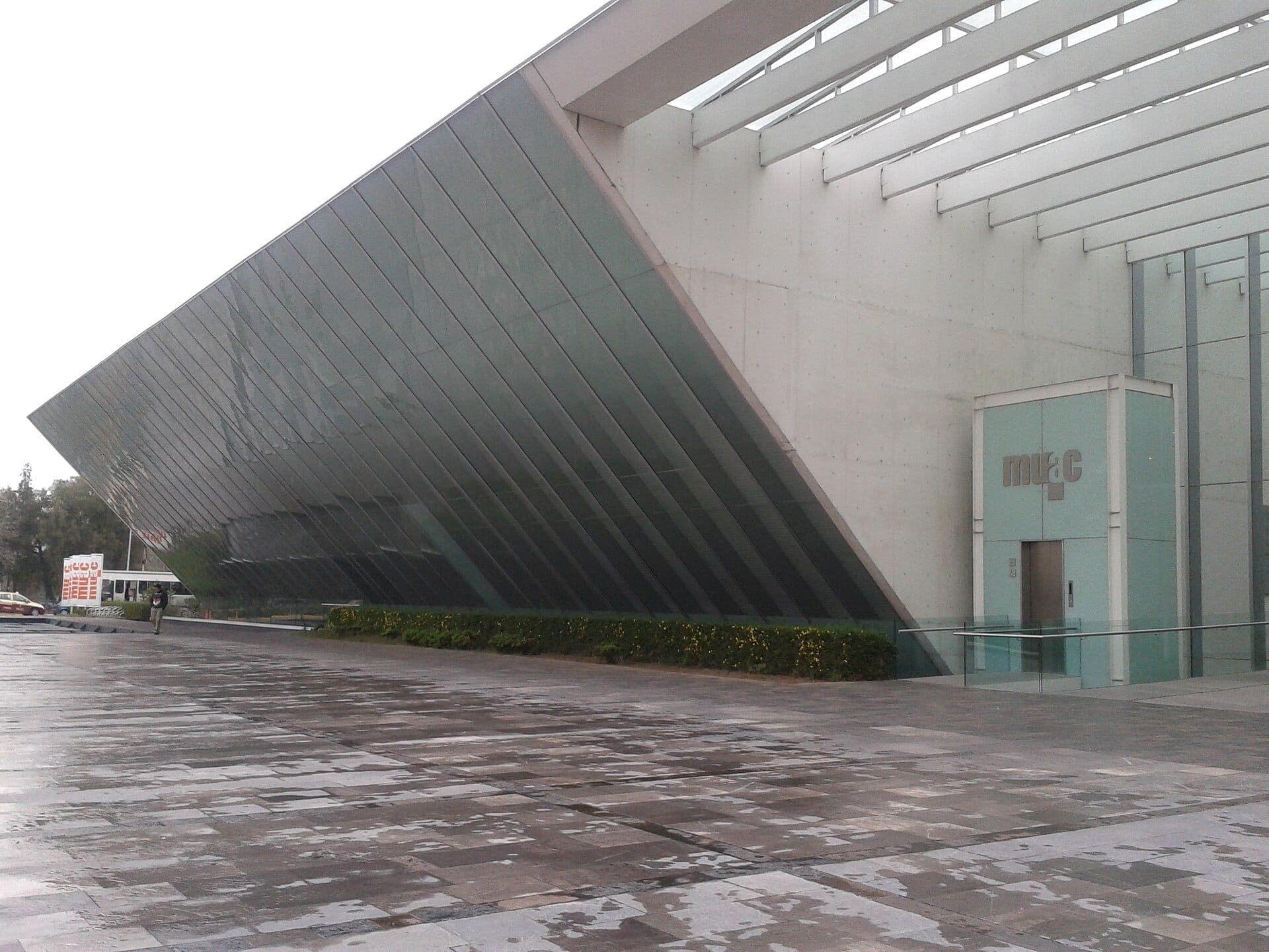 ด้านหน้าอาคารคอนกรีตและกระจกเป็นมุมของ Museo Universitario Arte Contemporáneo (MUAC) พร้อมเส้นสายสถาปัตยกรรมสมัยใหม่และลานหินกว้างด้านหน้า