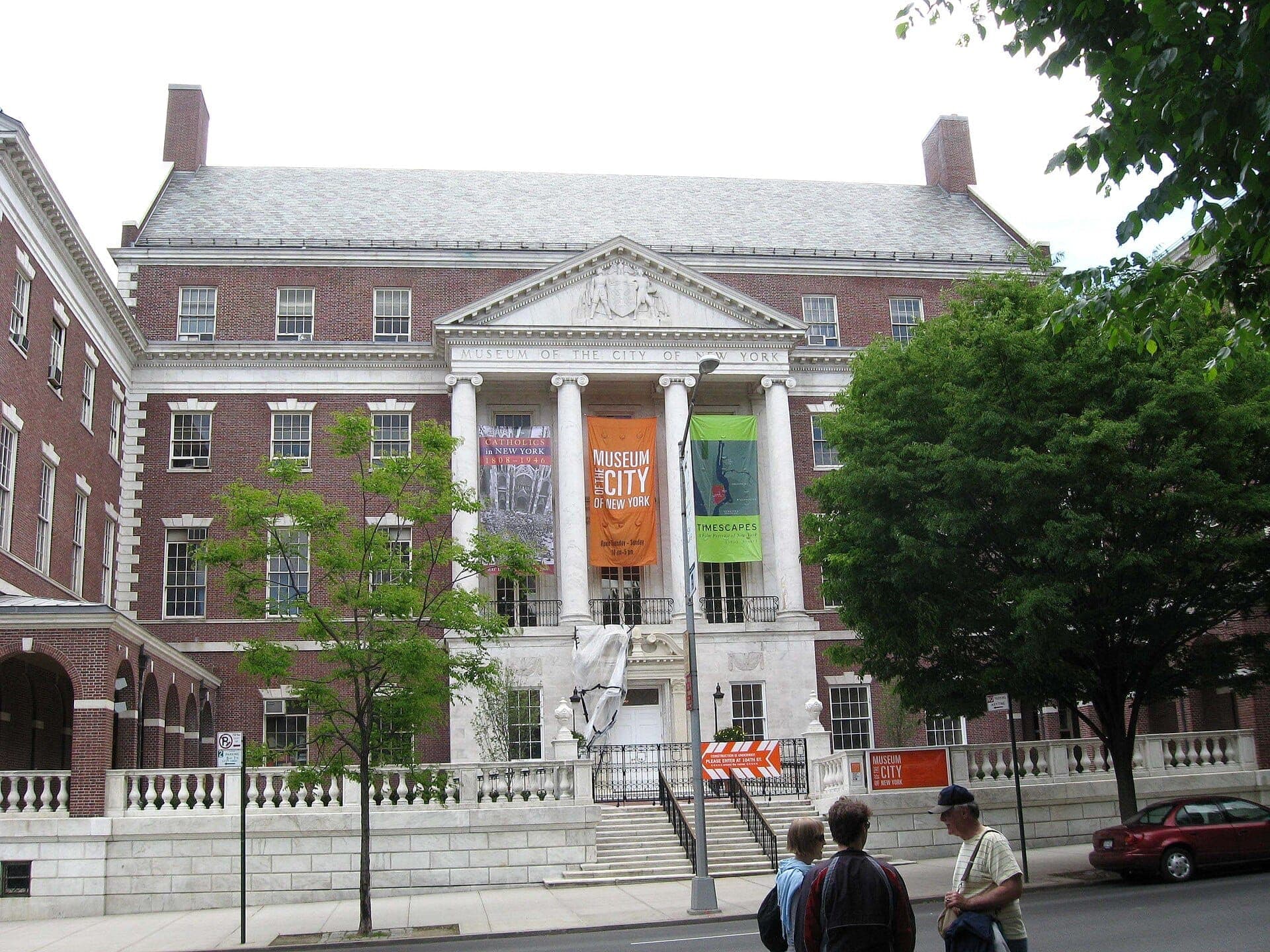 ด้านหน้าอาคารอิฐของ Museum of the City of New York พร้อมป้ายแบนเนอร์แขวนระหว่างเสาสีขาว ต้นไม้ และผู้คนบางส่วนบนทางเท้า
