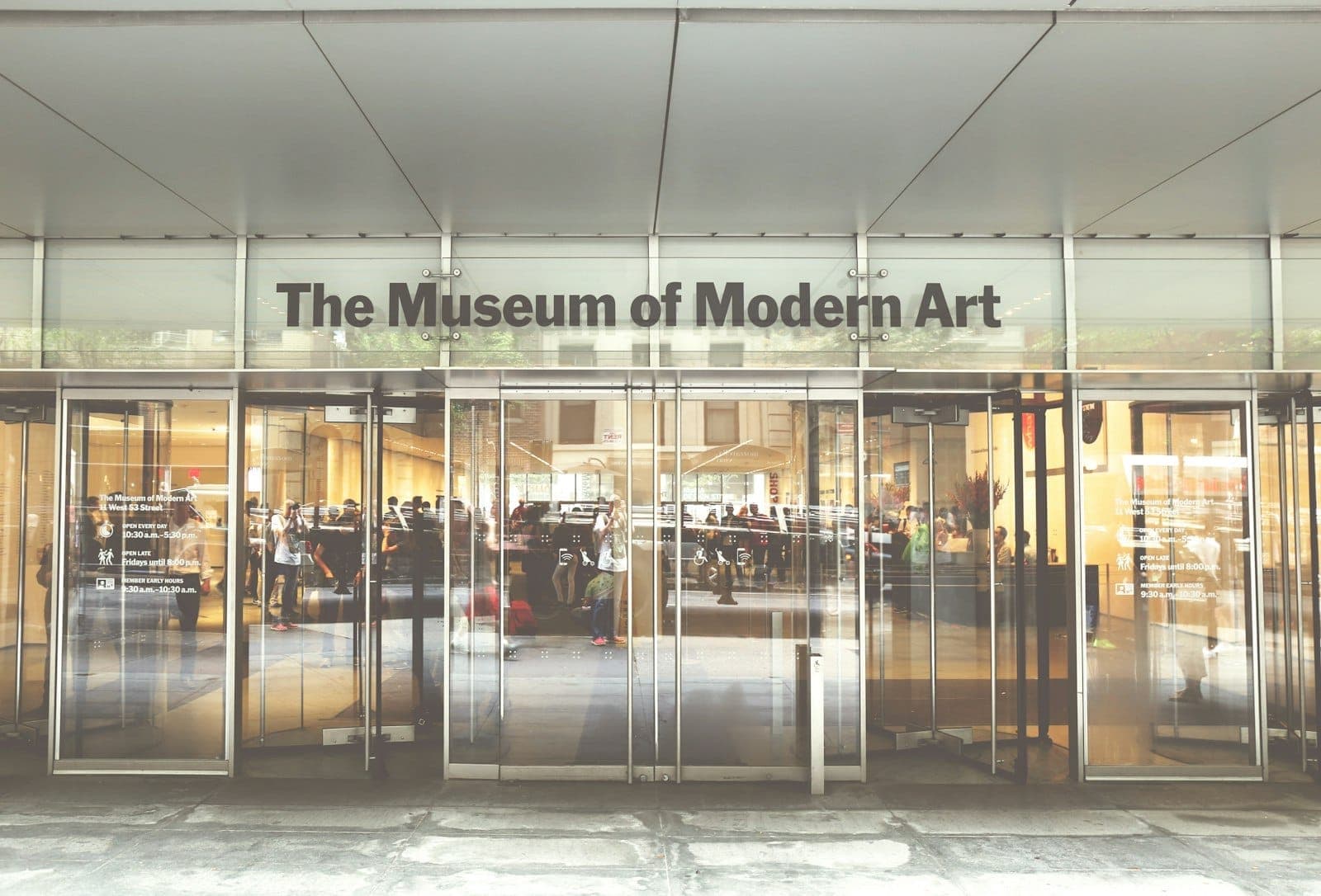 ทางเข้ากระจกของ The Museum of Modern Art พร้อมผู้เยี่ยมชมที่กำลังเข้าสู่ล็อบบี้ที่สว่างไสวและชื่อพิพิธภัณฑ์เหนือประตู