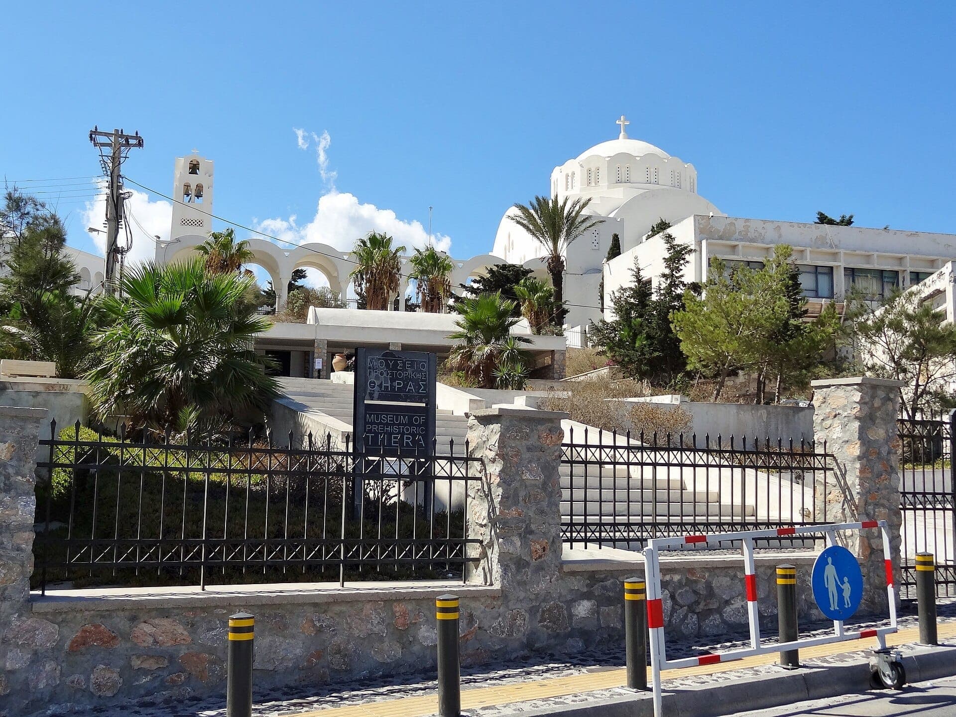 Straßenansicht des Museums für prähistorisches Thera in Fira, Santorini, mit seiner modernen weißen Fassade, Palmen und Eingangsschild an einem sonnigen Tag.