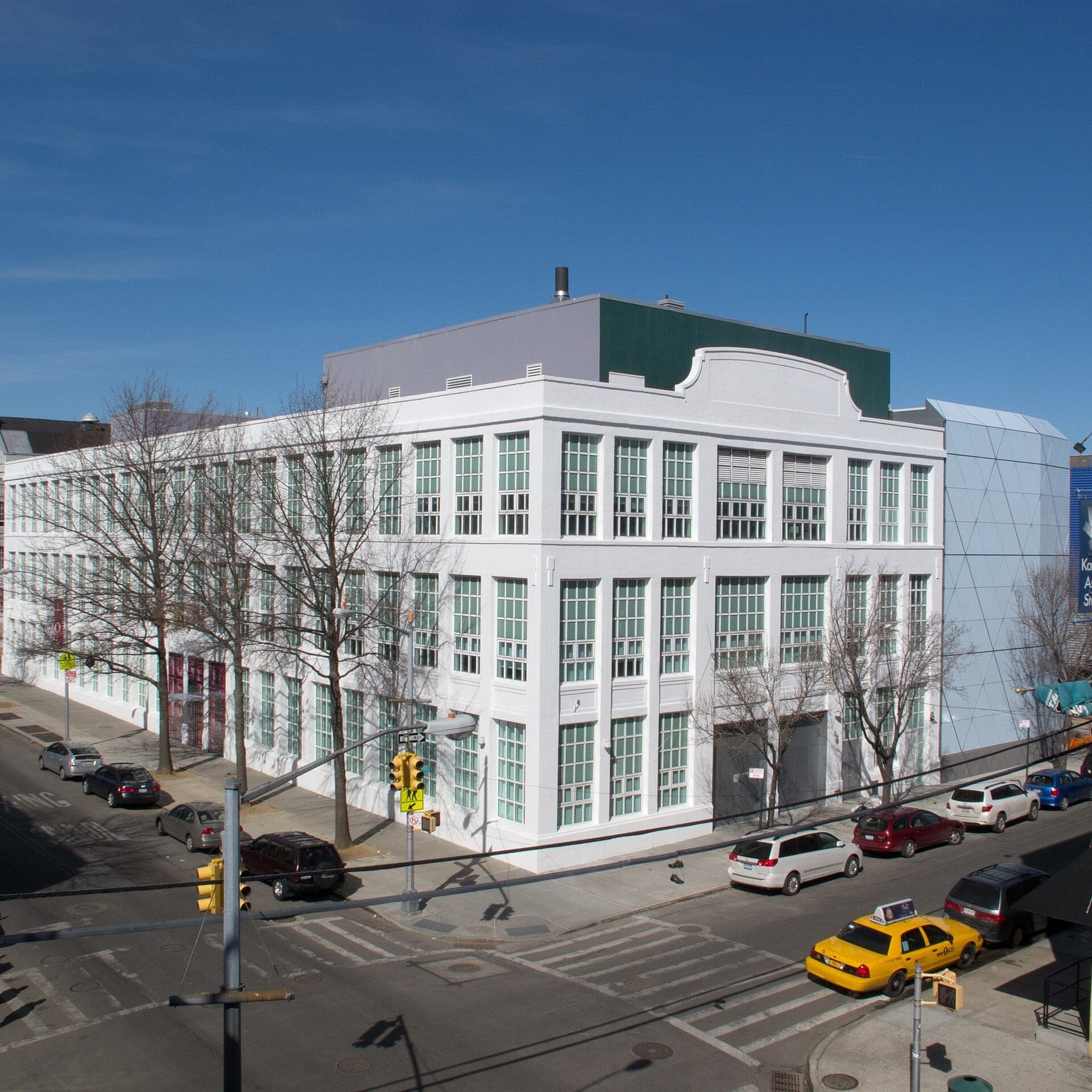 อาคารสีขาวที่มีหน้าต่างของ Museum of the Moving Image ที่มุมถนนในเมือง Astoria พร้อมรถยนต์และต้นไม้ไร้ใบเรียงรายตามถนน